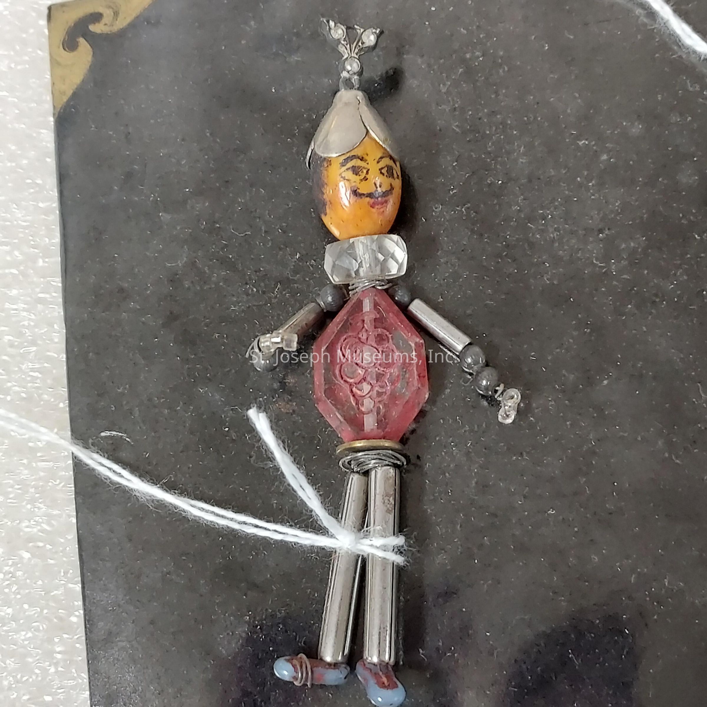 The image depicts a unique handmade pendant crafted to resemble a small human figure. The head is a painted bead with facial features, topped with a small hat-like piece. The torso consists of a large, faceted, reddish bead with designs, while the arms are made of cylindrical metal beads attached at the shoulders. The legs are similarly constructed using metal tubes, ending with small, blue beads suggesting shoes, and are secured with a piece of wire. The pendant is set against a dark background, highlighting its intricate design and the combination of different materials.