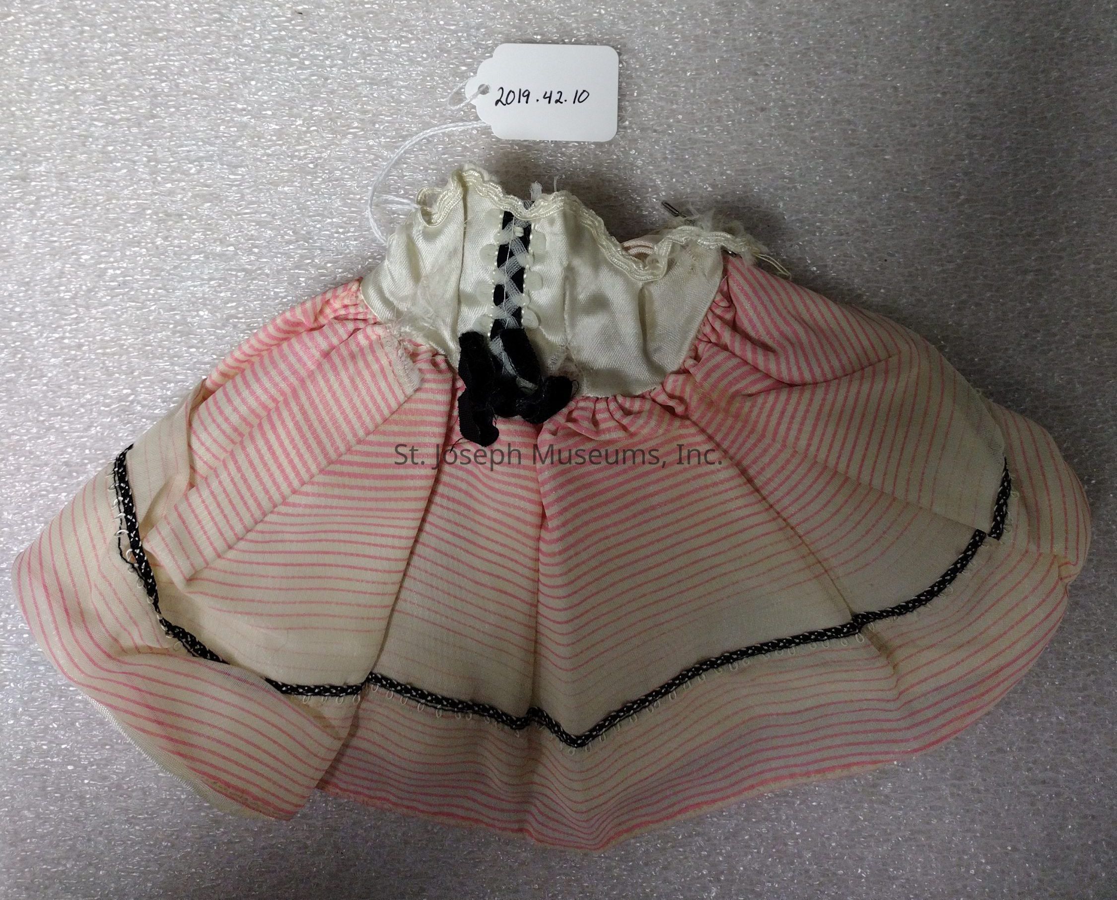 The image shows a small, vintage doll skirt with a delicate construction, featuring red and pink stripes on a light background, complemented by black lace trim along the hem and a gathered waist. The waistband is adorned with black ribbon lacing, and the fabric has a slightly sheer quality, adding to the antique charm. A tag labeled "2019.42.10" is attached to the garment, indicating its catalog or inventory number. The skirt is laid out on a textured, light background, suggesting it might be part of a collection or archival storage. The combination of colors and design elements suggests an old-fashioned style, characteristic of a certain period in doll fashion, and the construction appears detailed, showcasing skill in miniature garment making. The overall appearance gives a sense of nostalgia, evoking the elegance and style of a bygone era, possibly representing fashion trends from a specific historical period reflected in doll clothing.