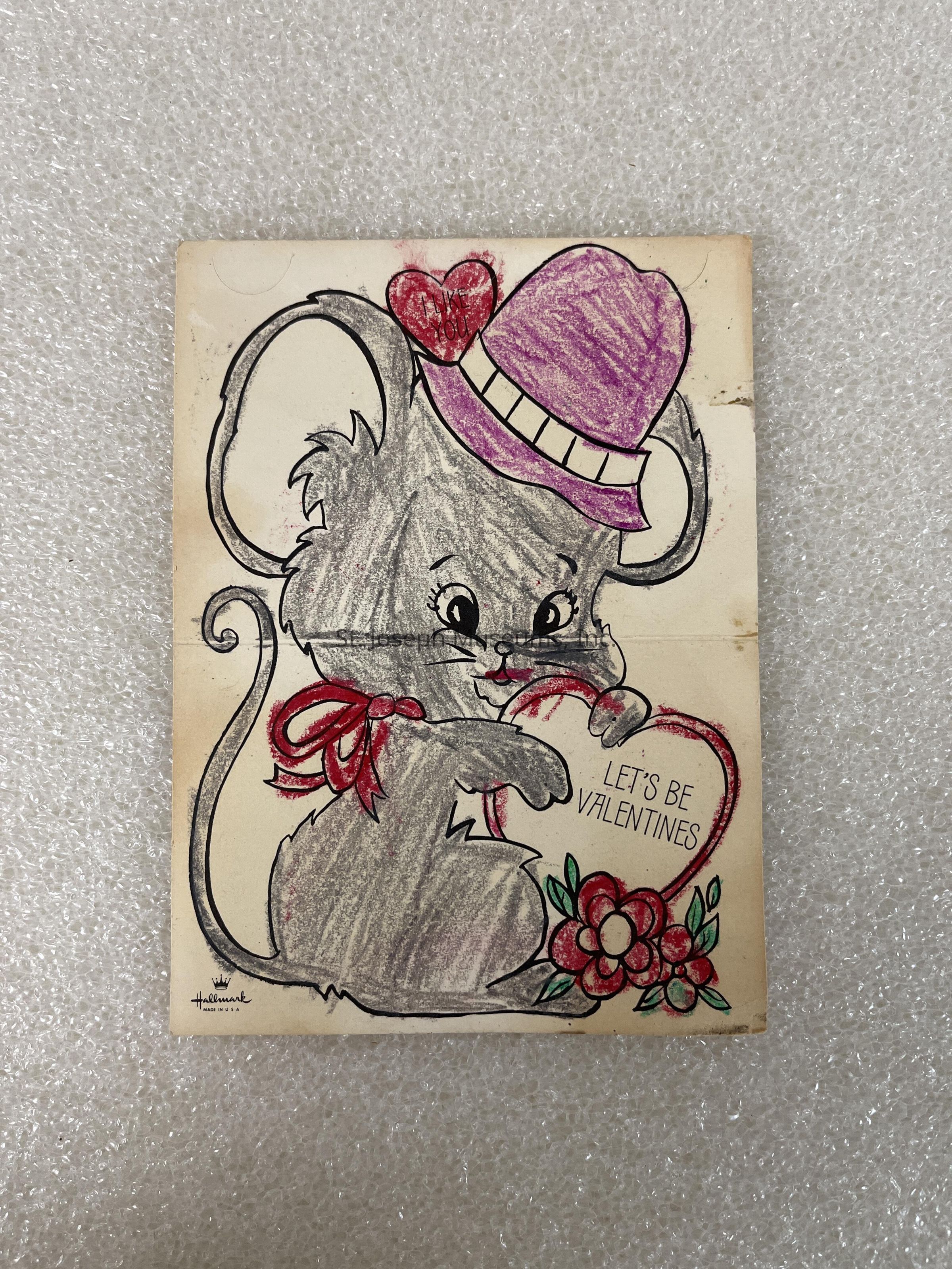 The image depicts a vintage-style Valentine card featuring an adorable cartoon mouse with large ears, who is wearing a purple hat adorned with a heart shape on top of its head. The mouse is playfully holding a heart-shaped object with the words “LET’S BE VALENTINES” written inside. Around the heart, there are bright and colorful flowers, adding a cheerful touch. The mouse's charming expression and the vibrant red and pink accents give the card a warm and nostalgic appeal. The Hallmark logo appears at the bottom, suggesting it is a classic design from the brand.