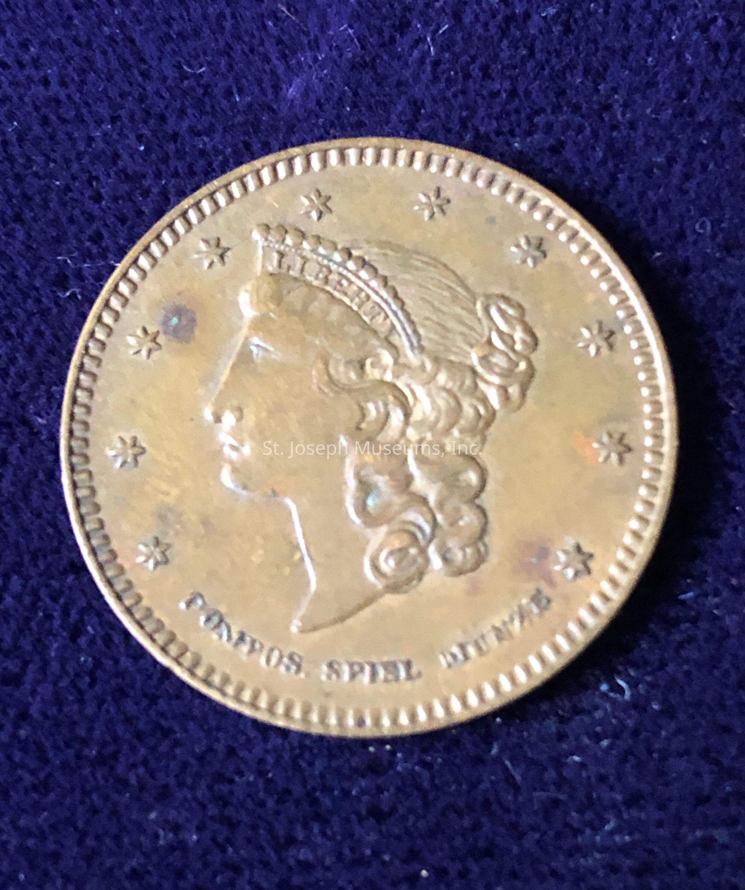 The coin displays a profile of a woman with detailed, curly hair and a tiara or headband across her head, suggesting a classical style or representation of a female figure. Encircling the portrait are several small stars, evenly spaced around the edge of the coin, contributing to its ornate design. Below the portrait, there is an inscription that reads "FORBES SPIEL MÜNZE," indicating a possible reference to the coin's origin, type, or use. The outer edge of the coin is decorated with a finely detailed border, which enhances the intricate craftsmanship of the piece. The overall appearance of the coin suggests it might be a commemorative or collectible item, rather than a standard currency for everyday transactions, given its artistic details and the specific text inscribed on it. The coin's metallic sheen is subtly captured by the lighting, highlighting the raised features of the woman's profile and the surrounding elements, thus