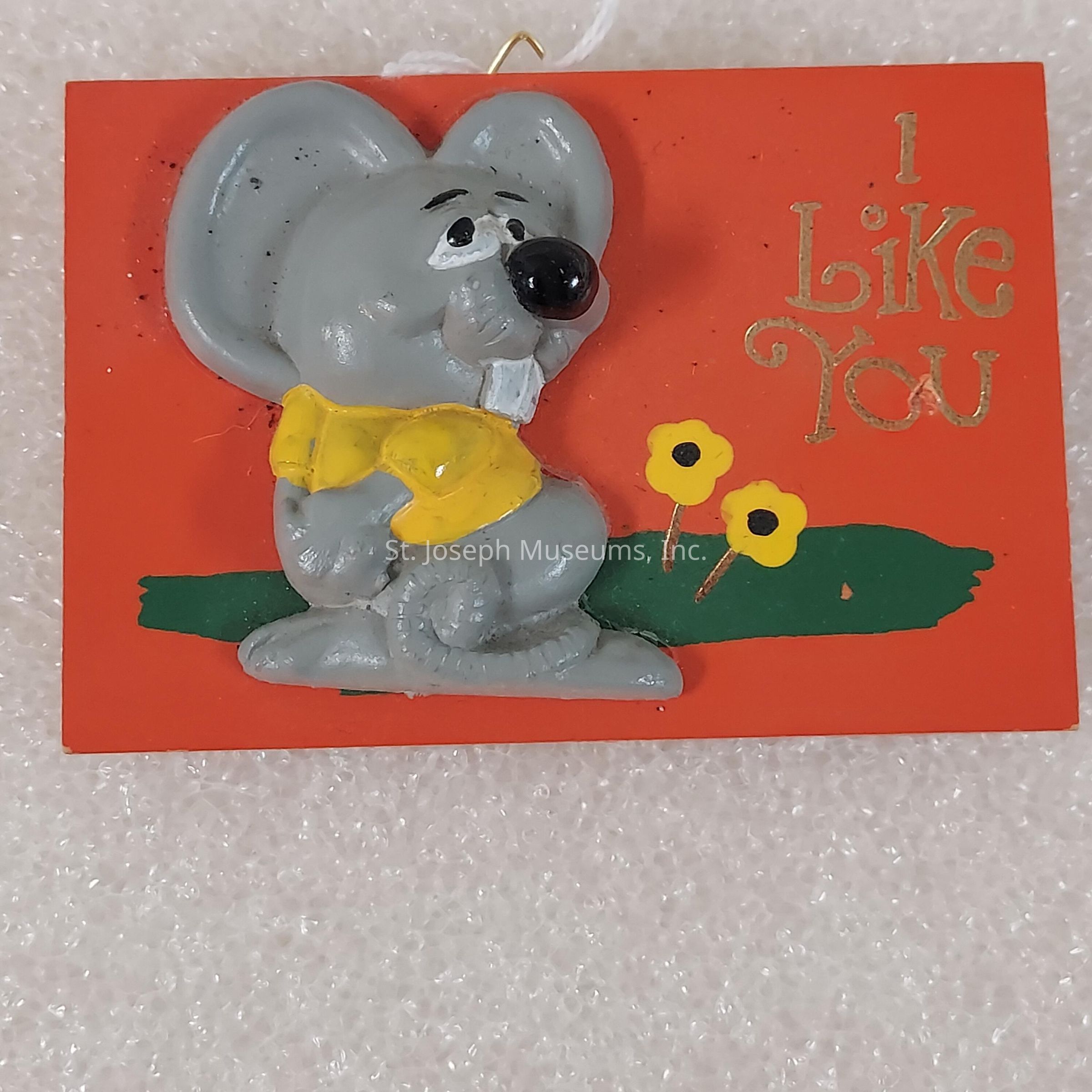The image features a playful decorative plaque with a cartoonish gray mouse donning a cheerful yellow bowtie. This adorable character is sitting on a grassy patch with two small yellow flowers depicted next to it. Above the mouse, the phrase "I Like You" is inscribed in yellow, stylized letters on the bright orange background of the plaque, adding a layer of warmth and friendliness to its overall appearance. The contrast between the vibrant orange background and the muted colors of the mouse and flowers creates a visually appealing effect. The textured surface on which the plaque rests further enhances its charm.