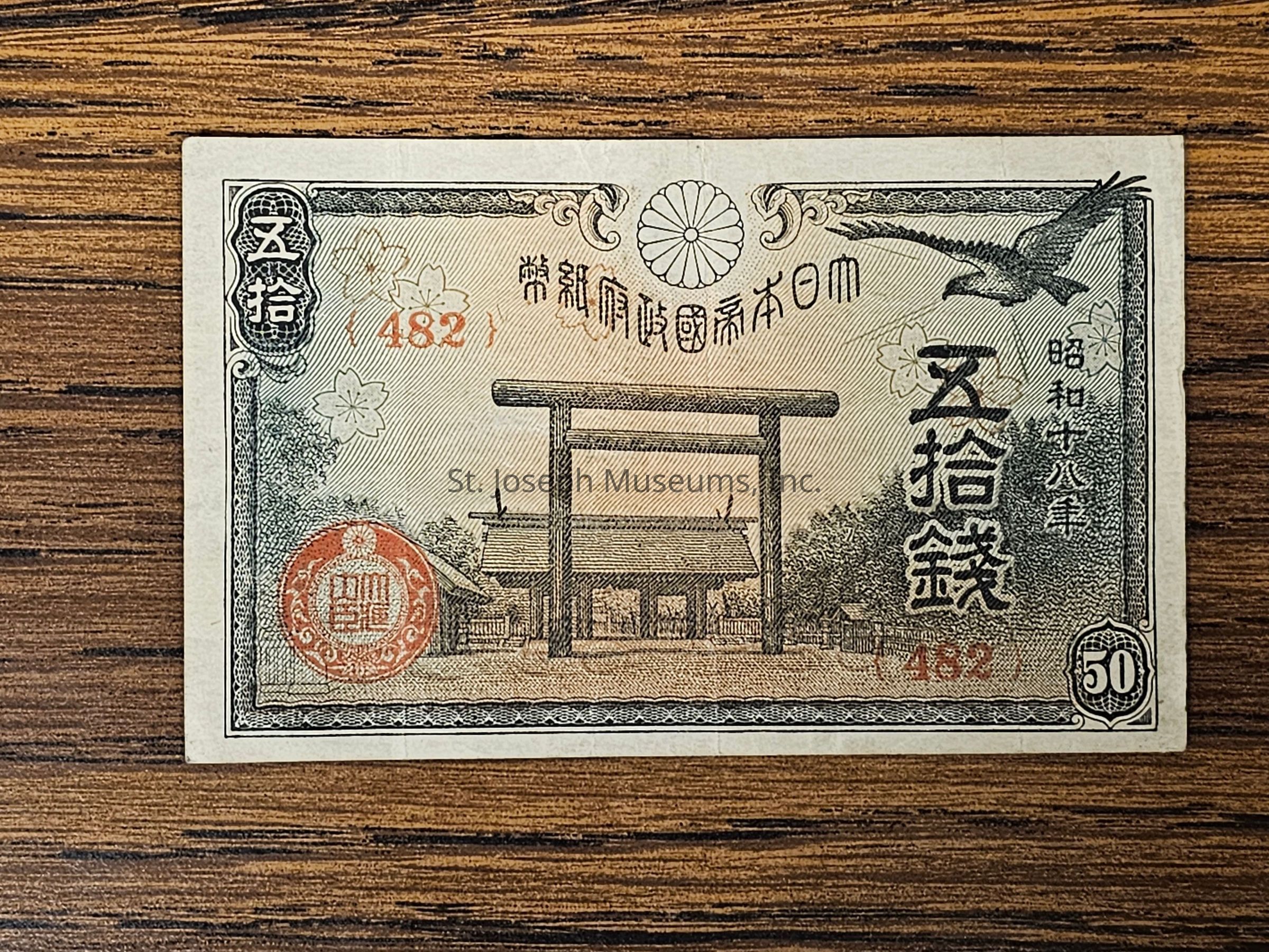 The image displays a vintage Japanese banknote with intricate designs and text in Japanese characters, including a denomination of 50 sen. It features a prominent Torii gate, a symbol of Shinto shrines, in the center, with decorative elements such as cherry blossoms, a crane in flight, and additional ornate patterns surrounding the scene. Various colors like black, red, and green are used to enhance its visual appeal, creating an aesthetically pleasing and richly detailed piece of currency art.