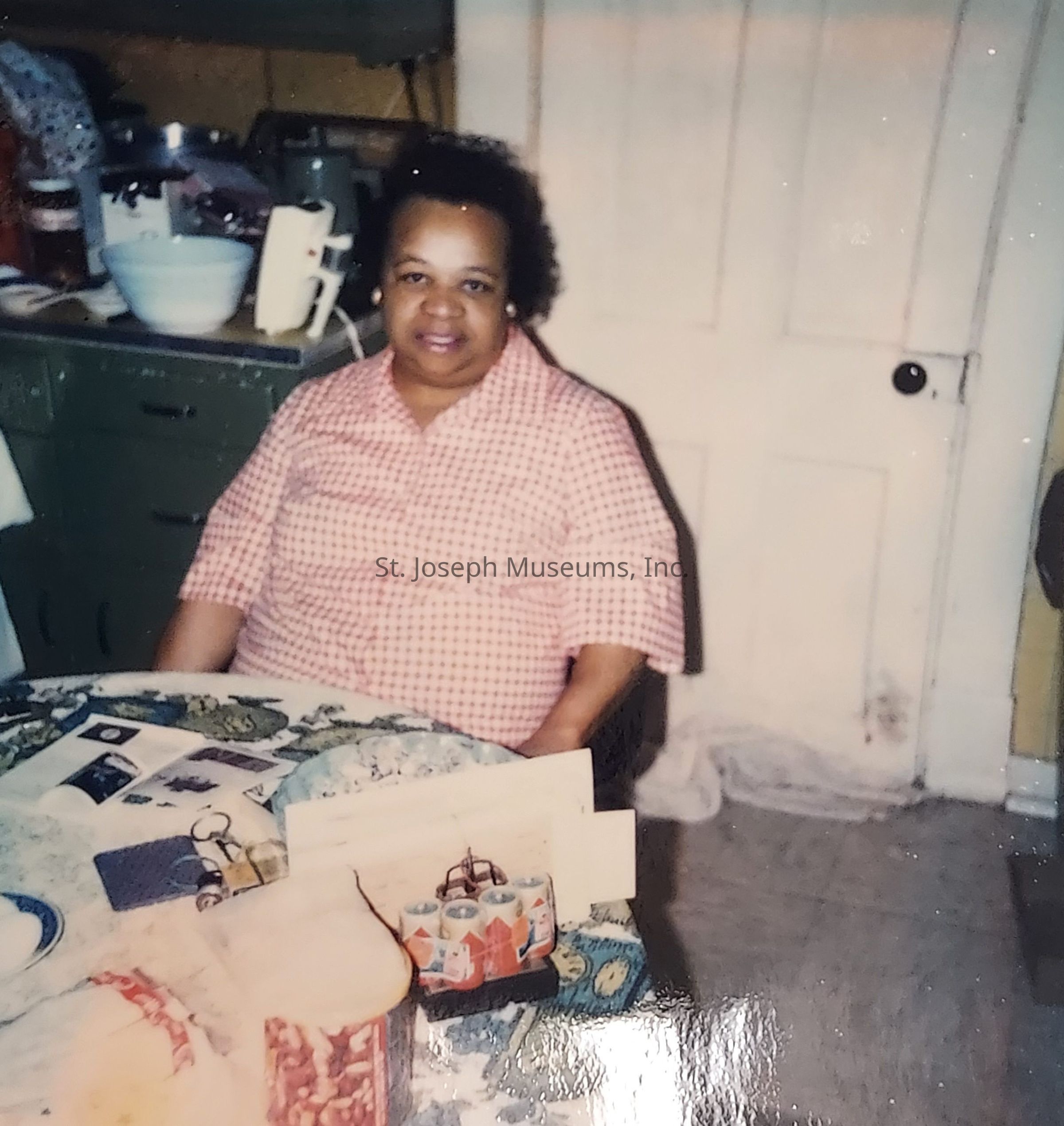 In the image, a person is seated at a table in a cozy kitchen with vintage decor, featuring a green cabinet and various kitchen items on the countertop, including a mixing bowl and a jar. The person is wearing a pink checkered shirt and smiling gently at the camera. The table is covered with a floral-patterned tablecloth and displays assorted items, such as cards and a small canister set. A white door with a dark handle is visible in the background, contributing to the warm and nostalgic atmosphere of the scene.