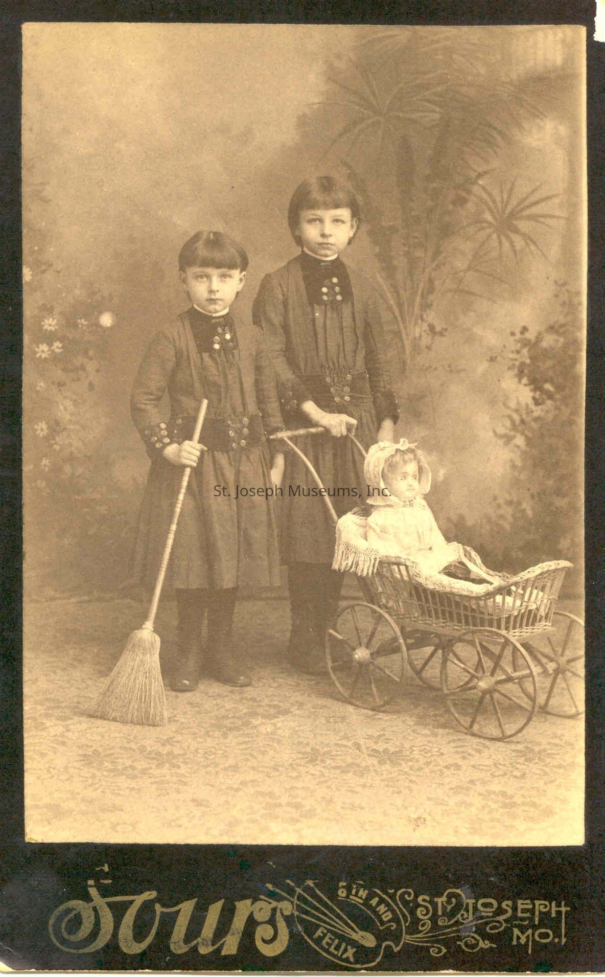 Caroline & Clara Zunkel with doll carriage, c. 1889: In the vintage sepia-toned photograph, two young children stand closely together, exuding an aura of early 20th-century charm. Both are dressed in matching dark dresses adorned with high collars, long sleeves, and large button embellishments, suggesting a formal occasion. The child on the left holds a broom, demonstrating a hint of domestic chore, while the slightly taller child stands beside them, gripping the handle of an old-fashioned wicker doll carriage. The carriage, intricately woven, cradles a doll wrapped in a delicately fringed blanket, its porcelain face framed by a lace bonnet. The backdrop, a studio setting, showcases soft, painted foliage with hints of palm leaves, adding a touch of elegance and warmth to the scene. The image, likely taken in St. Joseph, MO, as indicated by the decorative typography at the bottom, captures a poignant moment in time, blending elements of childhood innocence with the cultural and fashion norms of a bygone era.