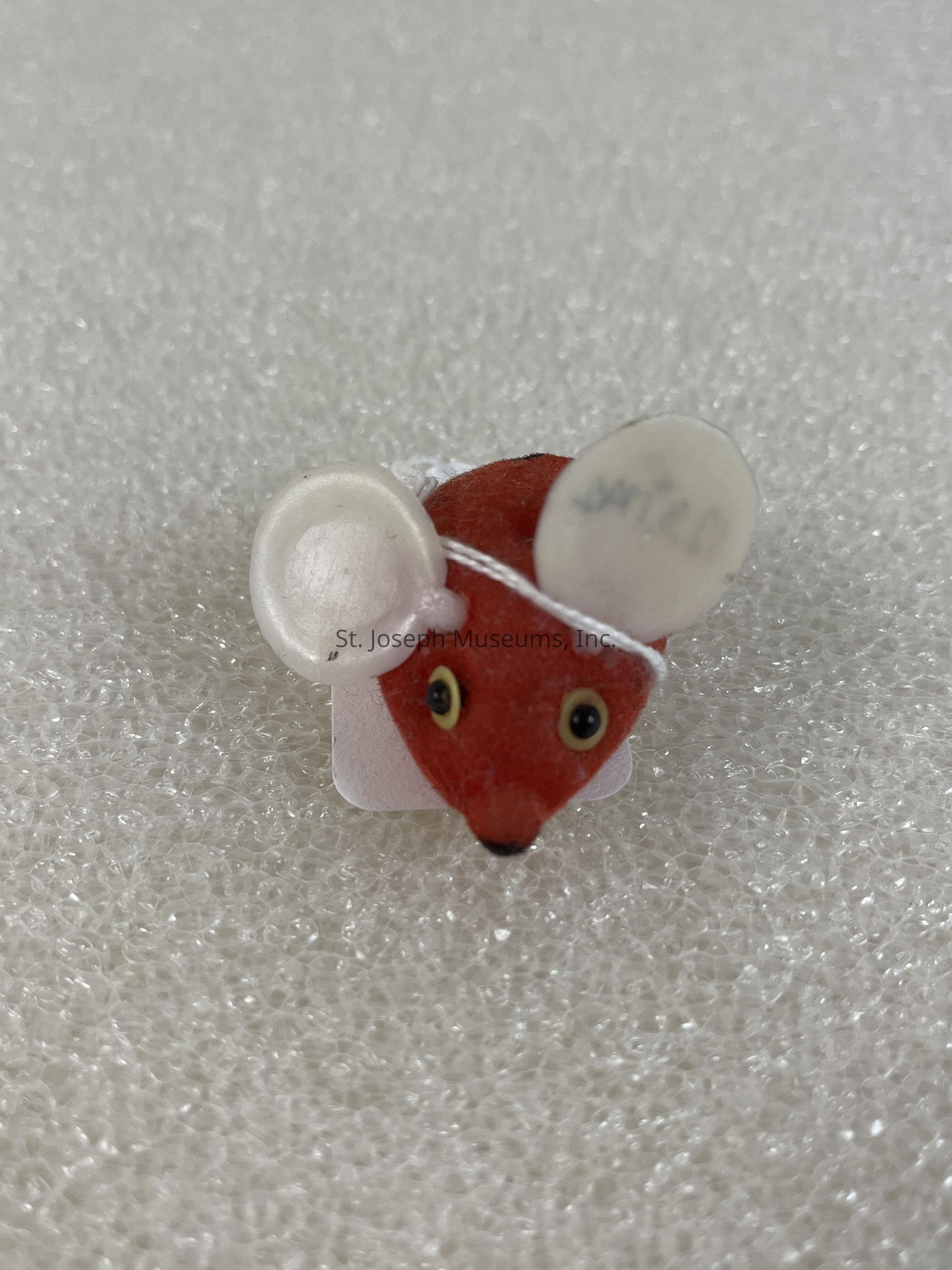 The image depicts a small red figurine resembling a fox, with bold yellow eyes and black pupils, positioned against a textured white background. The figurine is adorned with two white circular attachments that resemble ears, one of which has faint writing on it. The positioning of the fox and its round features suggest an artistic or whimsical design, emphasized by the bright color contrast between the red body and the white background. The intricate details like the expressive eyes and the added elements create a playful and charming representation, potentially appealing as a decorative or novelty item.