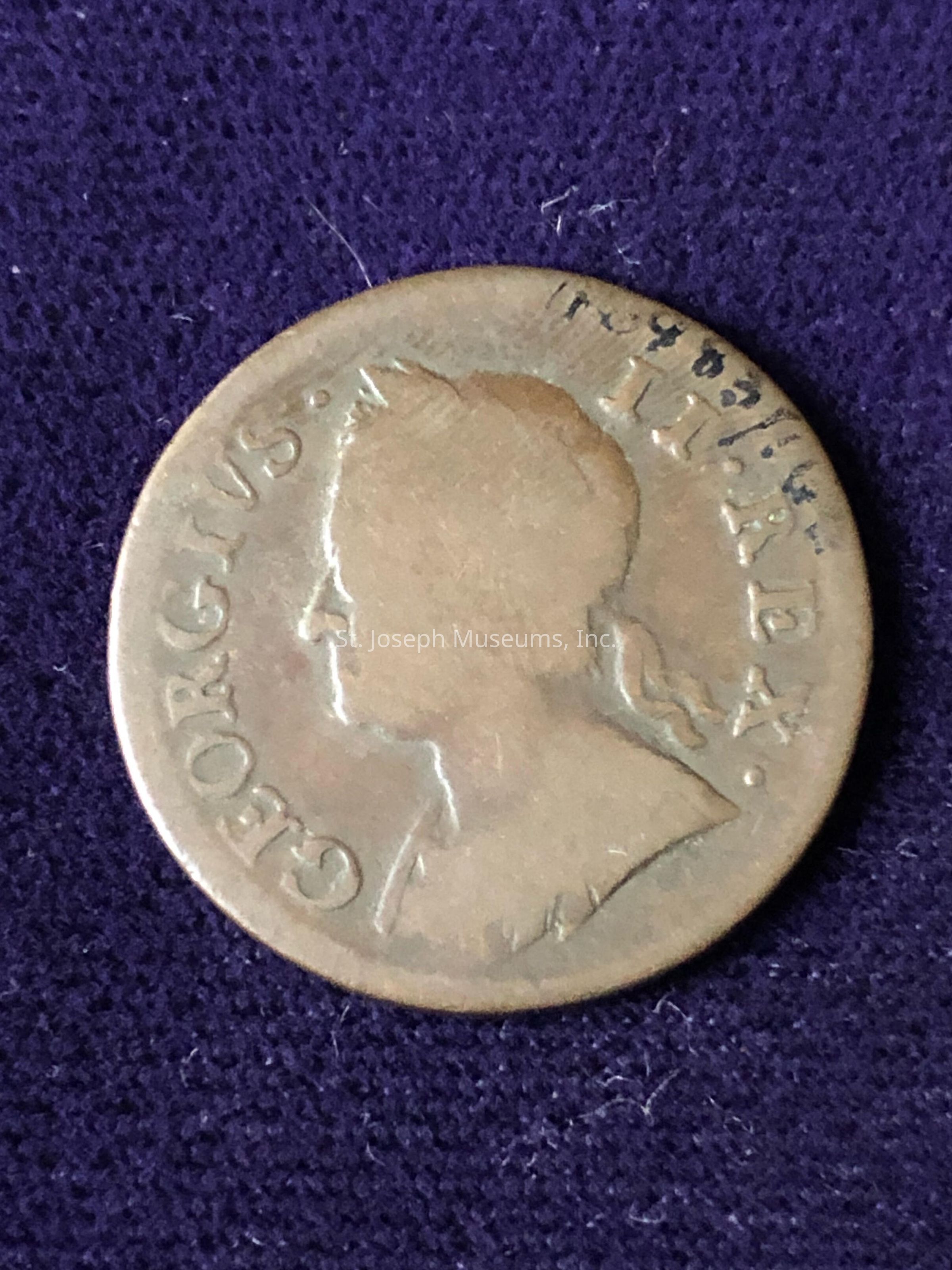 The coin appears to be made of a bronze-like material, showing some signs of age and wear. The obverse side, visible in the picture, displays an embossed profile of a person facing to the right, with the words "GEORGIVS" and other inscriptions sculpted around the portrait. The coin's surface exhibits a patina indicating its age. There is a distinct classical or vintage style to the design, typical of coins from past centuries. This image highlights the intricate details and artistry of coin minting, along with the coin's historical significance and potential value as a collector's item.