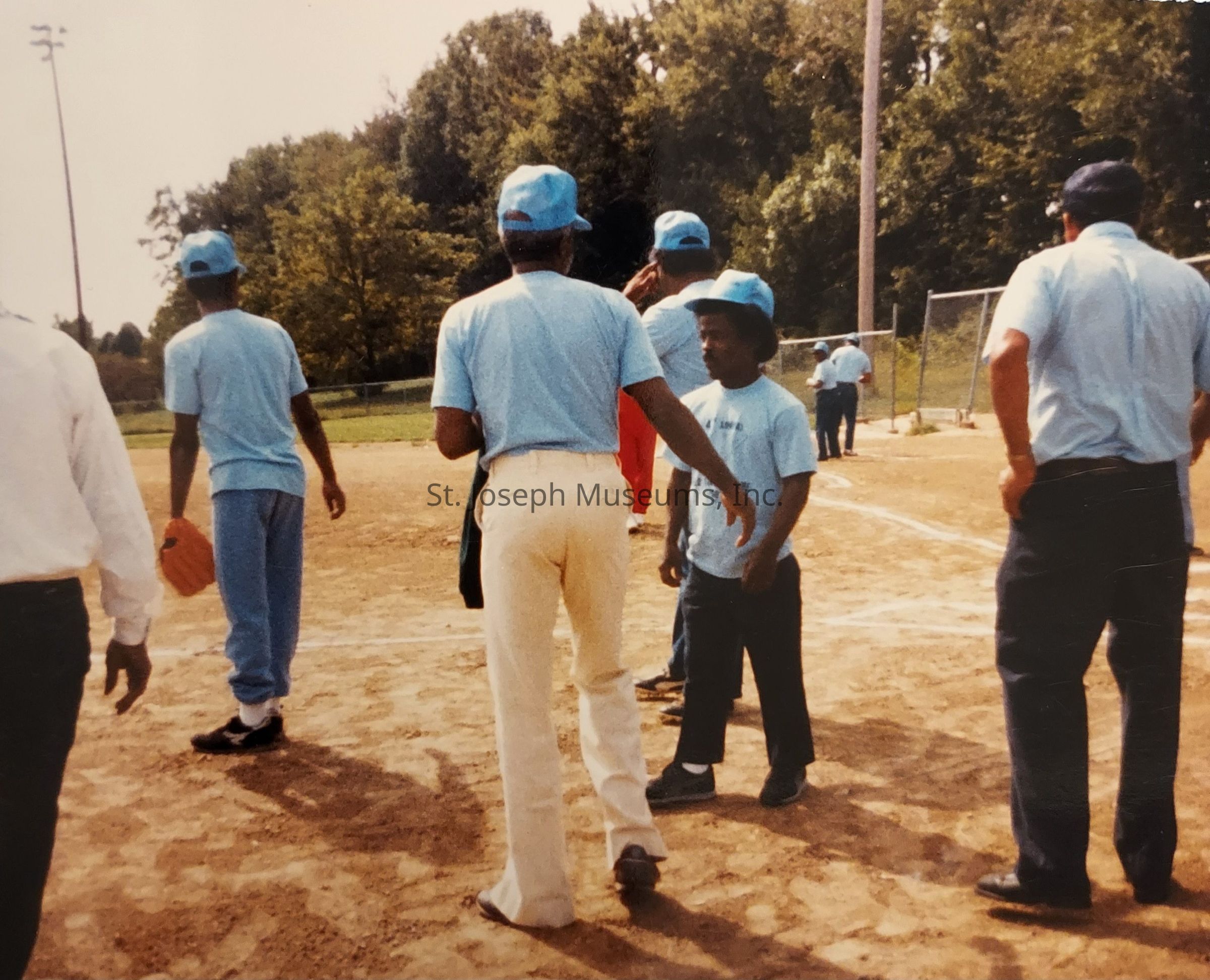 In this image, several people are gathered on a baseball field, all wearing light blue shirts and caps typical of a casual team uniform. The scene suggests a friendly or community baseball game, with individuals standing on the dirt terrain conversing and preparing for the activity. The background reveals lush green trees under a clear sky, indicating a sunny day suitable for outdoor sports. The players appear relaxed and engaged, with some holding baseball gloves. The casual attire and relaxed postures suggest a laid-back atmosphere, reflecting the camaraderie and teamwork often found in local sports gatherings.