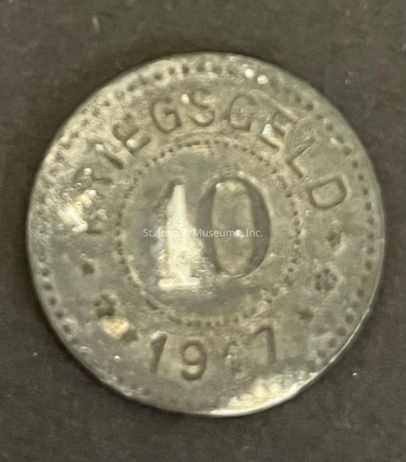 The image shows an old, worn coin with the word "KRIEGSGELD" prominently inscribed around the top edge, indicating that it is a type of emergency money issued during wartime, specifically referencing historical contexts such as World War periods in Germany. The coin, marked with the number "10" at its center, suggests its denomination, though the currency unit is not visible in the image. Below the denomination, the year "1917" is embossed on the coin, placing its origin during World War I. The coin's surface appears tarnished and shows signs of age, with a patina covering it, which is consistent with the materials commonly used for coins in the early 20th century. The design is simple, lacking intricate patterns or imagery, focusing primarily on its functional purpose as currency during a time of wartime instability. The background is a plain, dark surface, which contrasts with the coin, allowing its details to emerge more clearly, highlighting the historical artifact's util