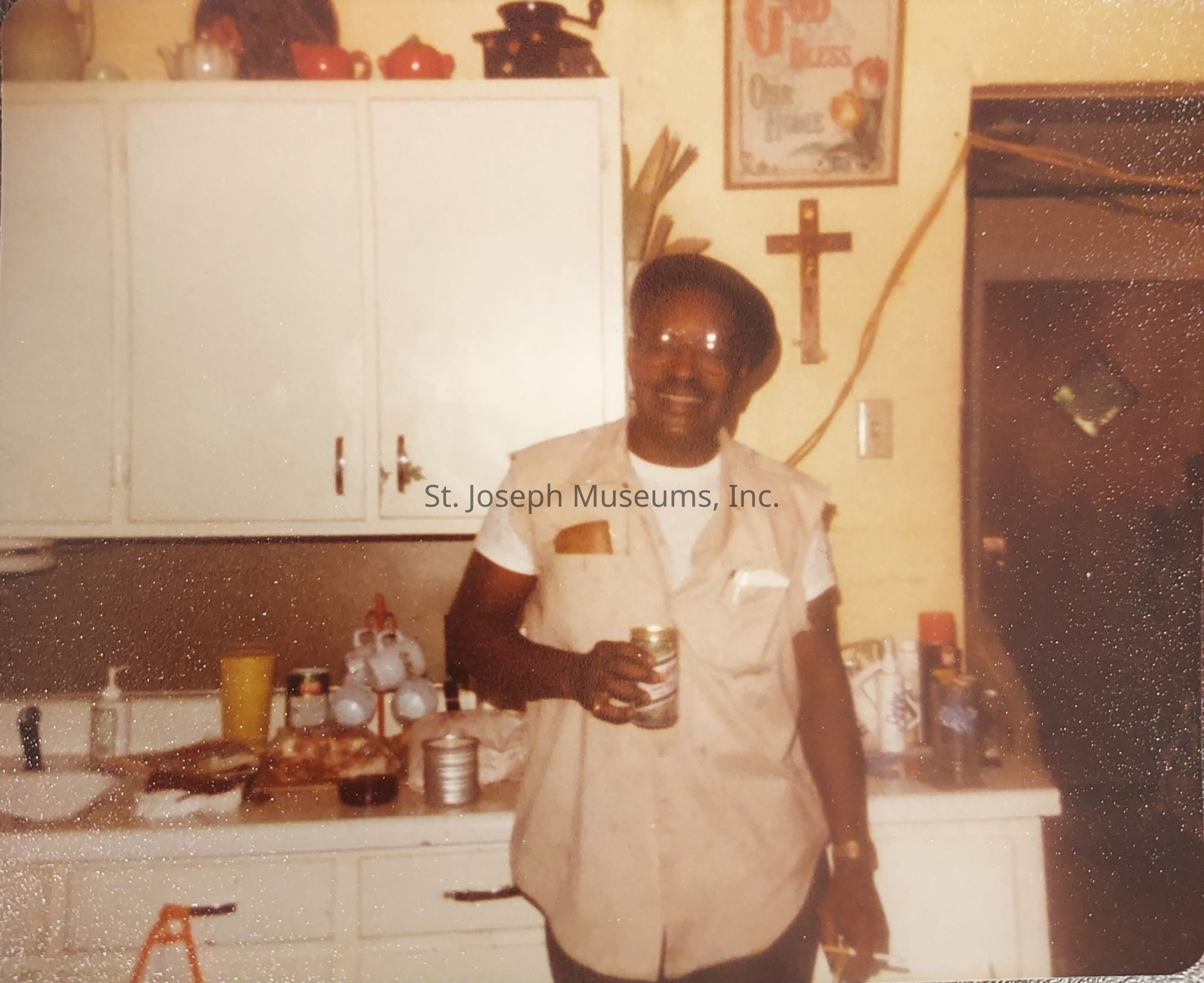 In this vintage photograph, an individual is standing in a warmly lit kitchen, holding a drink in one hand and a cigarette in the other, while wearing a beige vest over a white T-shirt and a dark beret; the background reveals a variety of items including kitchen cabinets adorned with colorful jars, a visible cross and picture frame on the wall, and a countertop cluttered with various kitchenware and products, indicating a casual, lived-in atmosphere.
