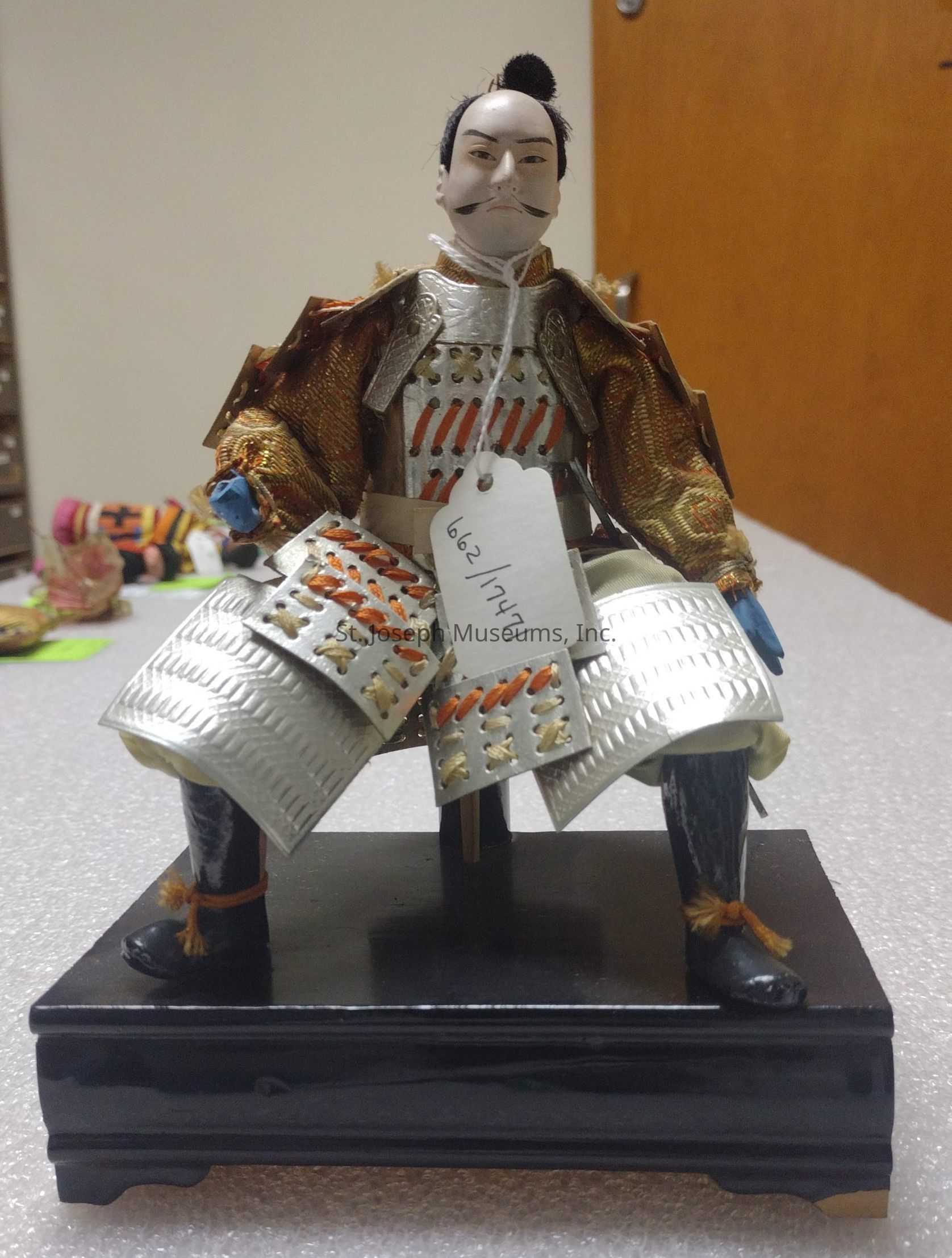 The image features a traditional Japanese doll, intricately dressed as a samurai warrior, sitting atop a black pedestal. The doll's attire is composed of detailed armor with white, silver, and orange elements that replicate the historical armor worn by samurai. The doll's stern face is highlighted with carefully painted facial features, including a thin mustache and a topknot hairstyle reminiscent of ancient Japan. A small tag is attached to the armor with numbers written on it, likely for cataloging purposes. The setting is a room with a blurred background, allowing the doll’s elaborate costume and graceful pose to capture attention.