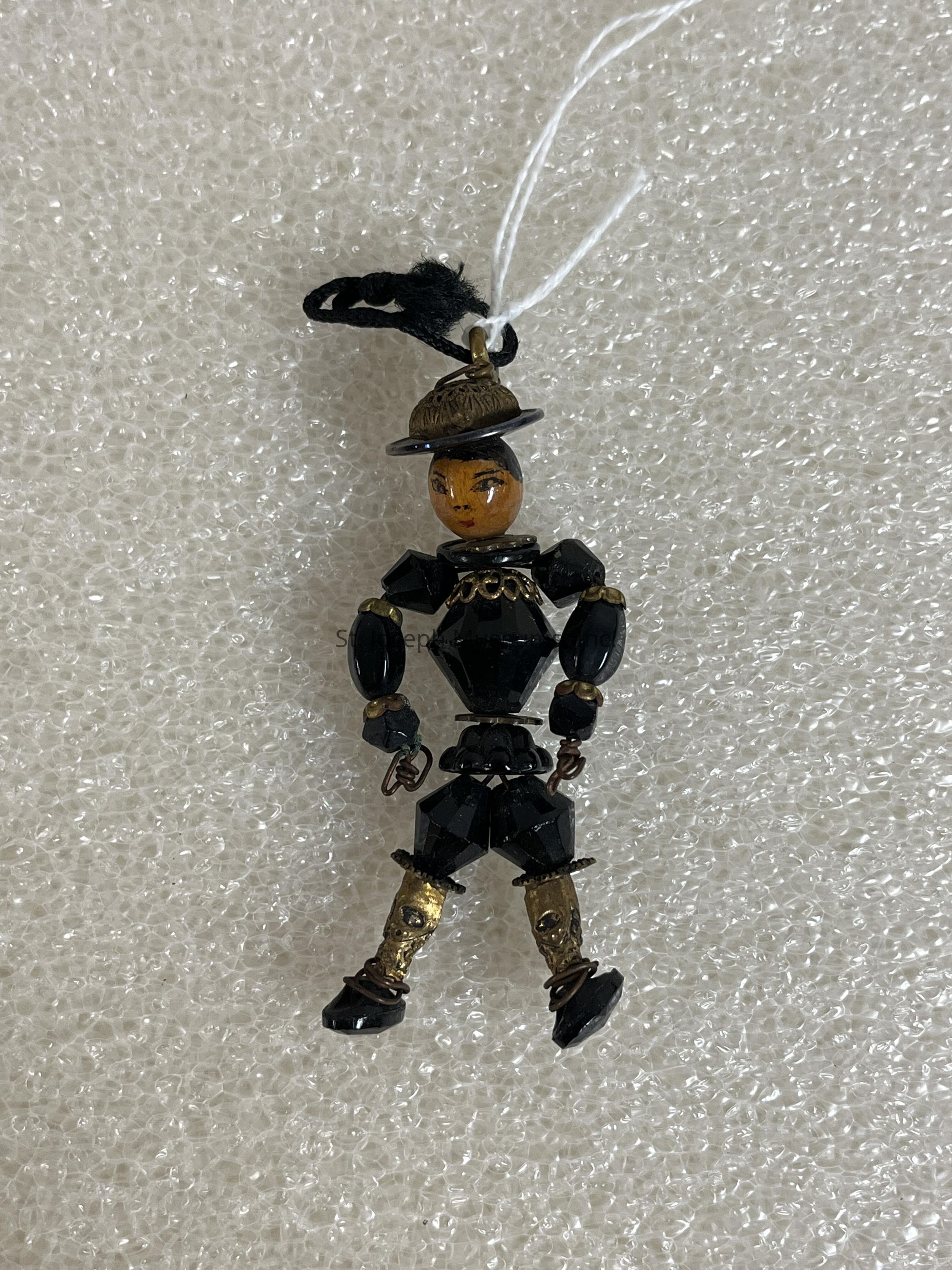 The image depicts a small, intricately crafted figurine resembling a puppet, with articulated limbs and a detailed, expressive face. The figurine is primarily black, with gold accents highlighting the joints at the shoulders, elbows, and knees. It wears a large black hat with a distinct brim and has a finely painted face that shows careful attention to detail. The puppet is strung with a white string attached to the top, suggesting it might serve as an ornament or decoration. The background features a textured, white surface resembling bubble wrap, which contrasts with the dark tones of the figurine, highlighting its sculptural features and adding depth to the overall composition. The artisan has skillfully balanced color and texture, creating a visually engaging miniature piece that exudes charm and character.