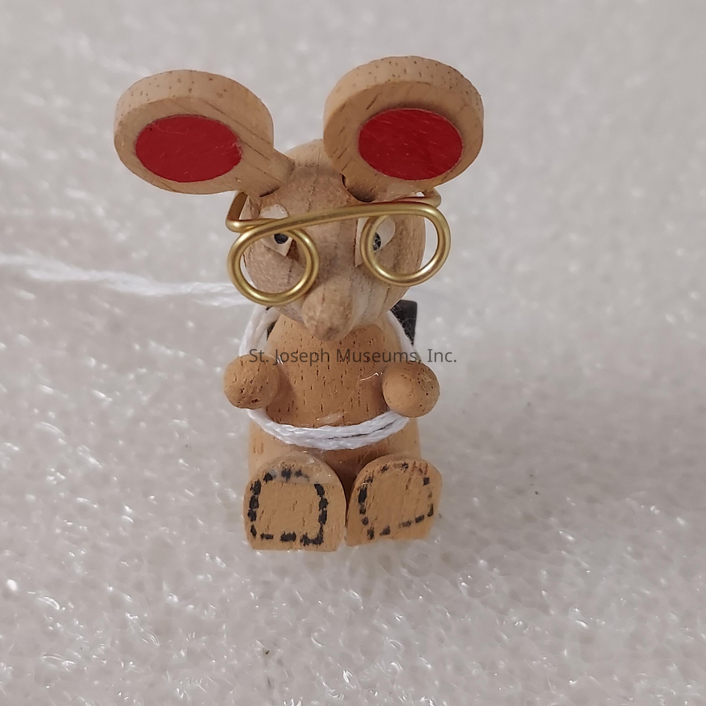 The image shows a small wooden figurine resembling a mouse, characterized by its whimsical design. The mouse features exaggerated round ears painted red, and it is wearing wire glasses crafted from thin gold-colored wire. The figurine is intricately carved, with details like small paws and feet. A thin white string is wrapped around its body, giving an impression of a scarf or some form of decoration. The mouse is set against a textured background, which appears to be a type of white foamy material, adding contrast to the earthy tones of the figurine, enhancing its playful and artistic depiction.