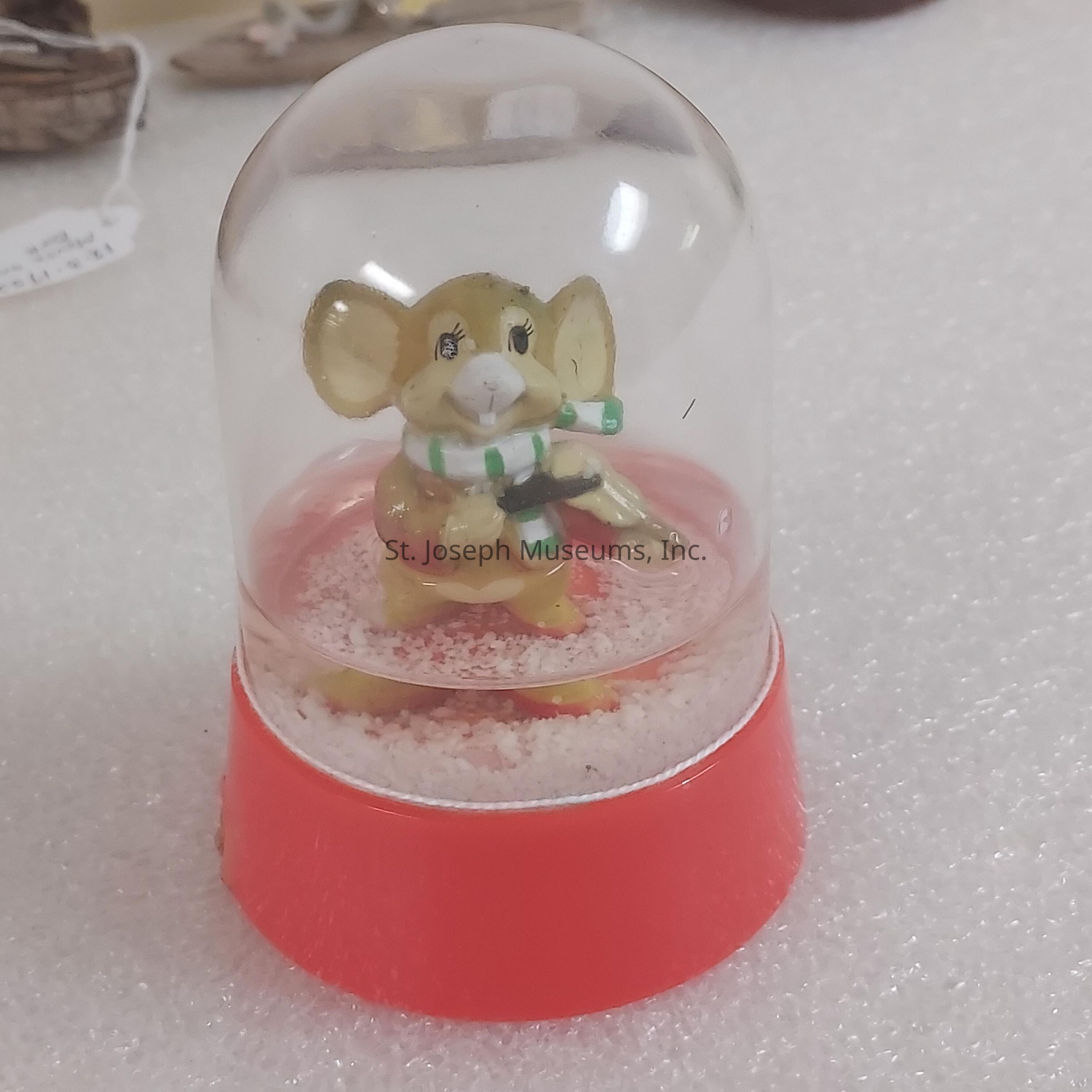The image shows a small snow globe featuring a whimsical figurine of a mouse inside; the mouse is wearing a green and white striped scarf and holding a musical instrument that resembles a guitar, creating a festive appearance. The mouse appears cheerful with wide eyes and prominent ears, capturing an air of joy and music within the tiny confines of this glass dome. The base of the snow globe is bright red, complementing the colorful interior figure. Around the mouse is a light sprinkling of faux snow, enhancing the wintry scene inside the globe. The background, though blurred, suggests a setting such as a store or a display area, where various decorative items might be showcased. The overall mood conveyed by the image is one of whimsy and holiday cheer, bringing a smile to those who happen upon this delightful object, making it a charming piece for seasonal decoration or gift-giving, capturing the spirit of celebration in a small, self-contained world.