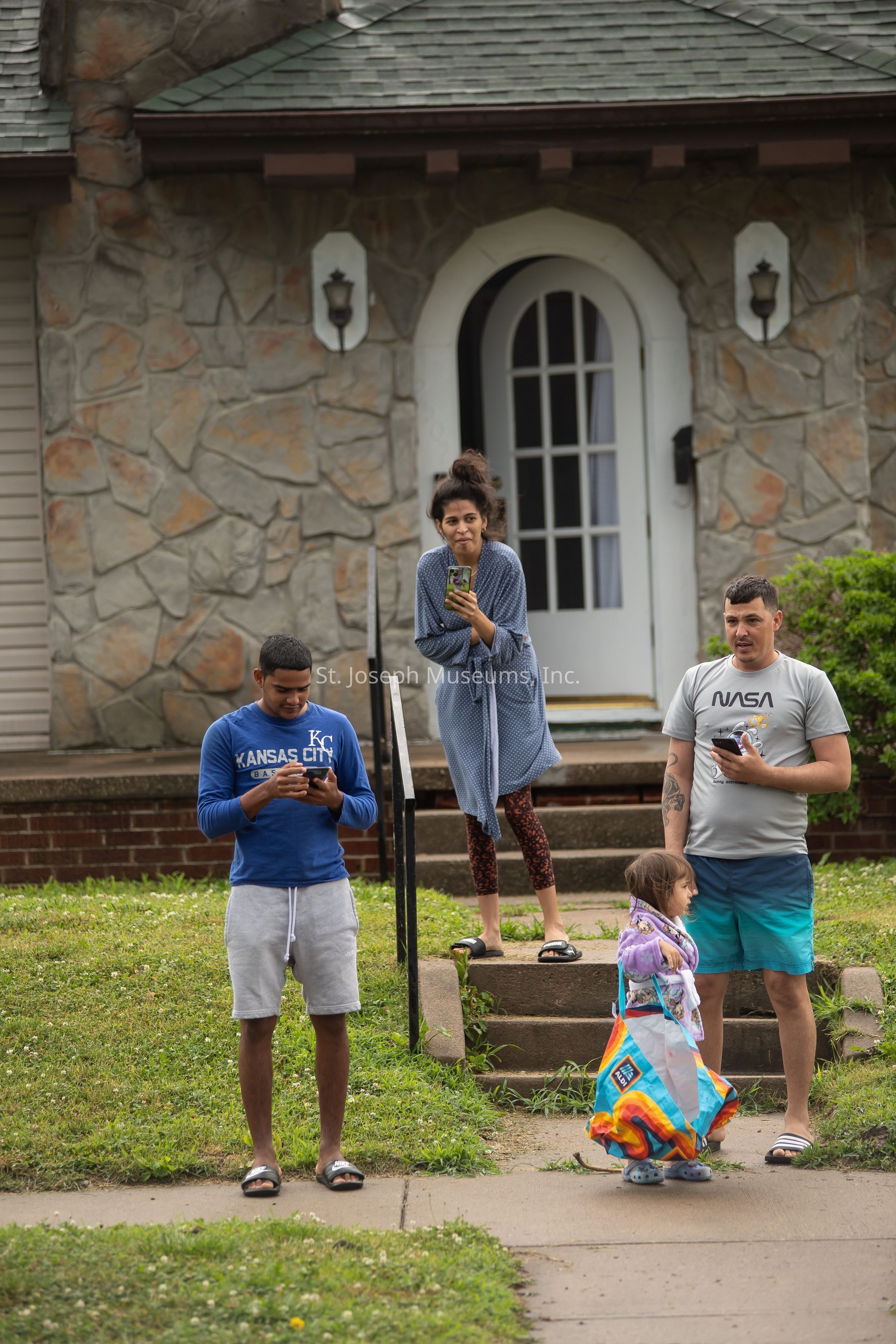 The image shows four people standing outdoors in front of a house with a stone facade and a white door. A man in a blue "Kansas City" sweatshirt is focused on his phone, standing on the grass. A woman in a blue robe and patterned leggings stands on the steps, holding a phone and smiling. Another man wearing a NASA t-shirt stands nearby, also looking at his phone. A young girl, wrapped in a colorful blanket and wearing purple shoes, stands in front of the man in the t-shirt. The scene conveys a casual moment, possibly depicting family or friends engaged in a lighthearted interaction.