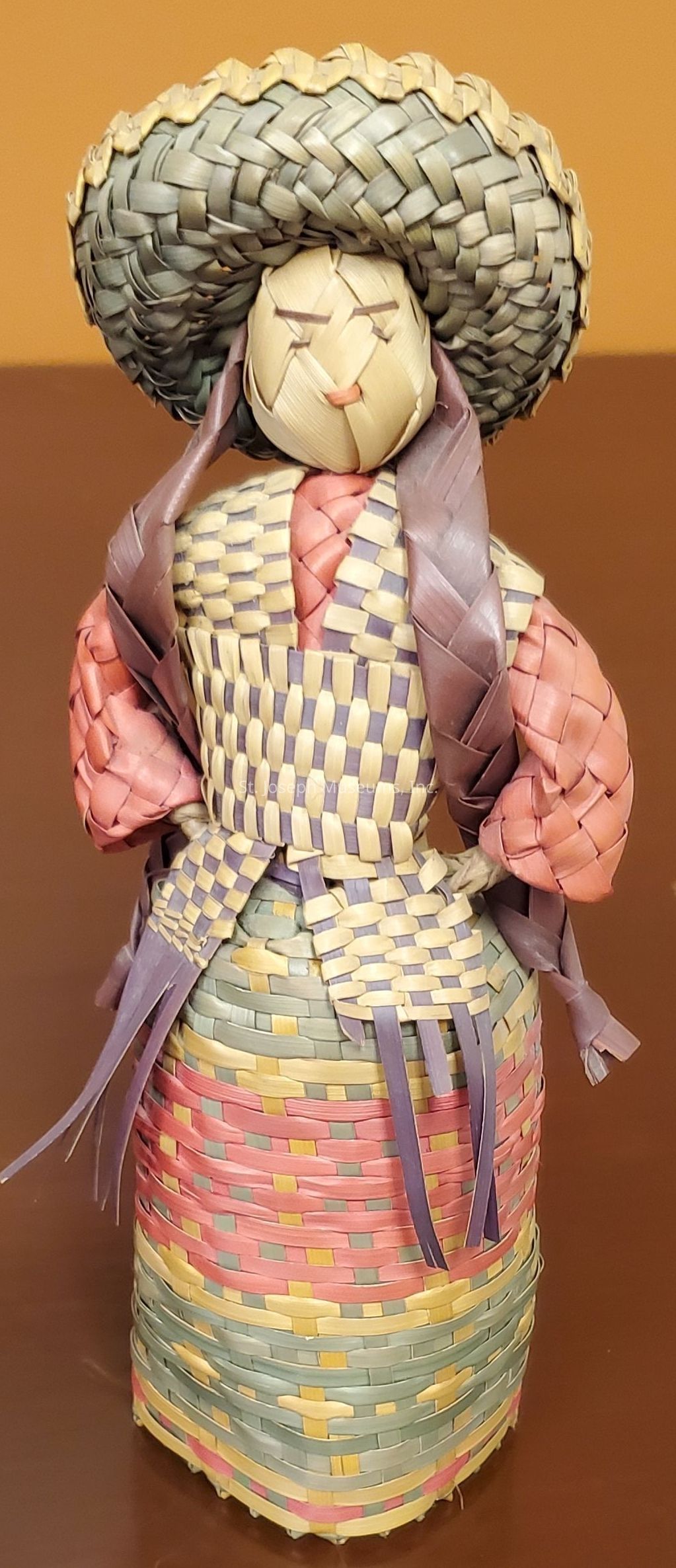 The image shows a handcrafted doll made entirely out of intricately woven straw and reed strands, featuring various shades of natural, muted colors such as pastel pink, lavender, cream, and grayish-green. The doll is elegantly dressed, with a woven wide-brimmed hat and what appears to be a traditional outfit, consisting of a multi-colored, checkered top and a long woven skirt with horizontal bands. Her arms are styled in pink tones, and she has additional braids draping down. The craftsmanship highlights attention to texture and structure, with the straw woven in tight, precise patterns that offer a sense of both fragility and durability. The doll's face is simple and abstract, lacking detailed features, which contributes to its charm and rustic aesthetic. Overall, the piece embodies a fusion of cultural artistry and traditional craft, suggesting that it might be an artisan's representation of cultural heritage or a handcrafted souvenir.