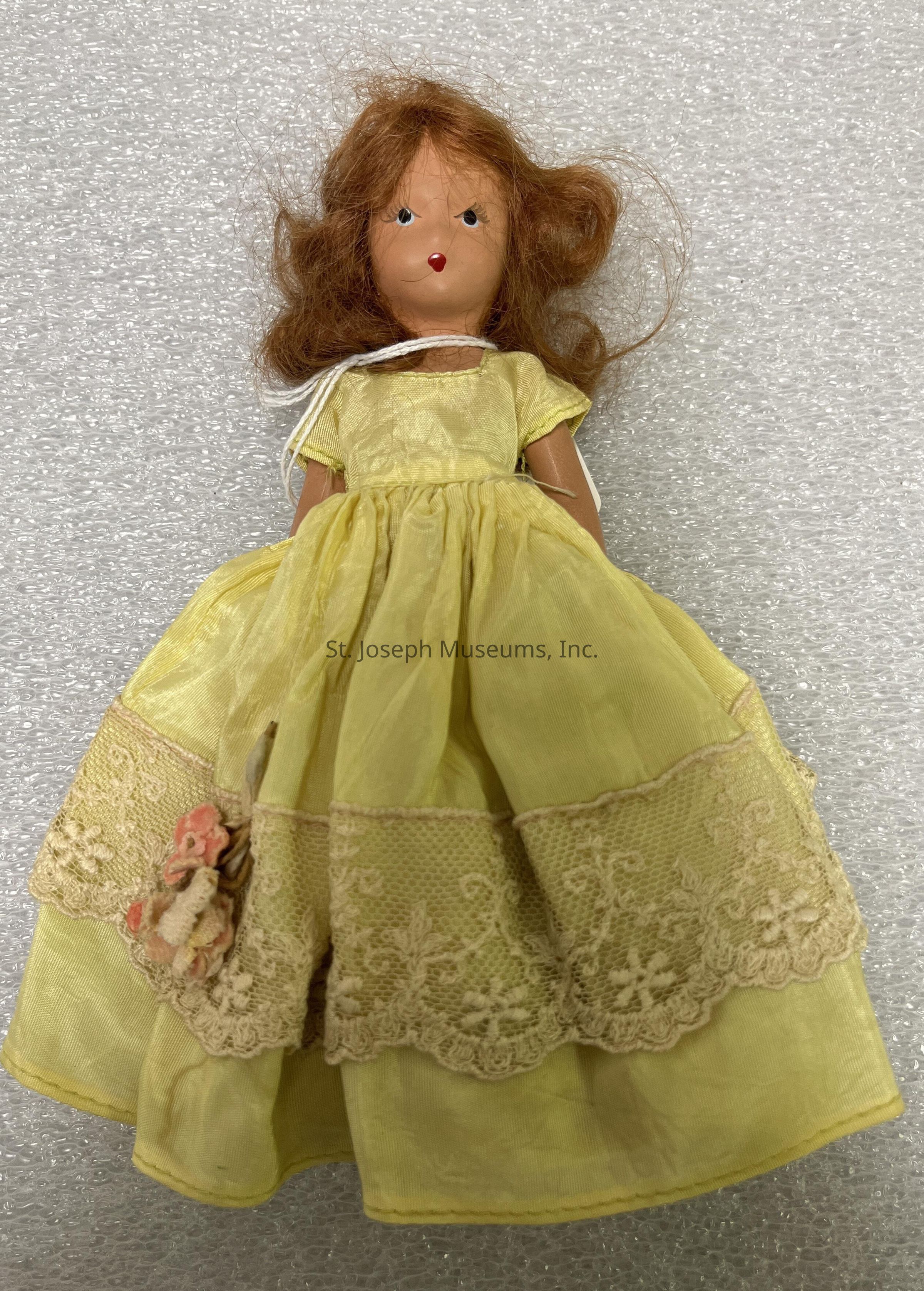 The image features a vintage doll with a serene facial expression, having vibrant red hair that is styled loosely and cascades around her face. She is dressed in an elegant yellow gown adorned with intricate lace detailing and floral decorations. The gown appears to have a soft, flowing texture, enhancing the doll's delicate and refined appearance. She is placed against a backdrop of a textured white surface, possibly bubble wrap, which adds a contrasting modern element to the scene. The doll's classic and antique design suggests it may hold sentimental value or be considered a collectible item.