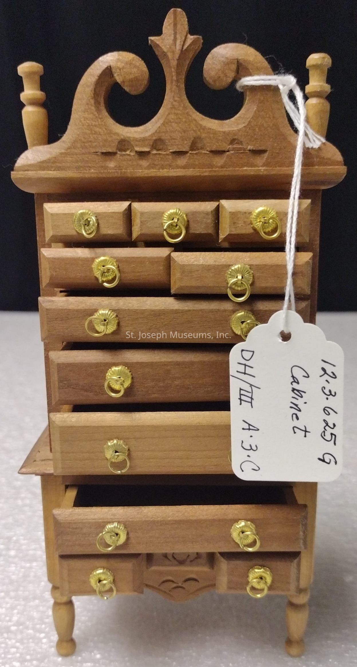 The image shows a small, intricately carved wooden cabinet with multiple drawers featuring gold-colored handles. The cabinet has an ornate top with scrollwork detailing and is topped by two small finials. It rests on four turned legs, and one of the lower drawers is slightly open. Attached to the cabinet is a white tag with handwritten markings, displaying what appears to be an identification code: "12.3.6259" along with the word "Cabinet" and a reference "DH/III A.3.C." The background is a simple, dark backdrop which highlights the craftsmanship and details of the miniature piece of furniture.