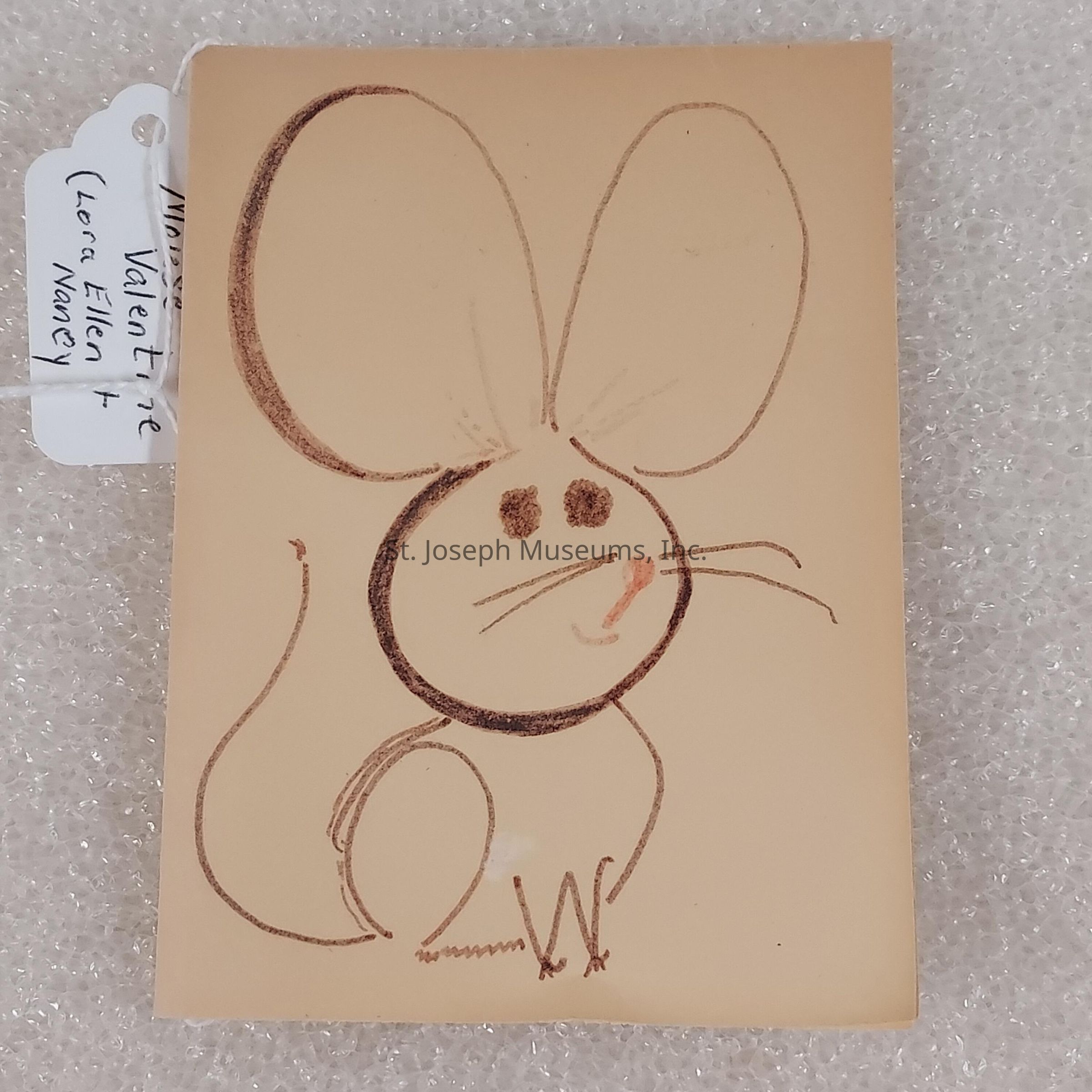 The image displays a simple, charming illustration of a mouse drawn on a light beige piece of paper. The mouse is depicted with large, oversized ears and prominent eyes, adding to its whimsical appearance. This straightforward line drawing uses soft brown and pink hues to outline its features, such as the round body, curved tail, and long whiskers. Below the paper, there is a textured, white bubble wrap surface visible. To the left of the main drawing, a small tag is attached with a string, showing handwritten notes. This composition exudes a playful and quaint charm, evoking the simplicity and warmth of handmade art or a child's drawing.