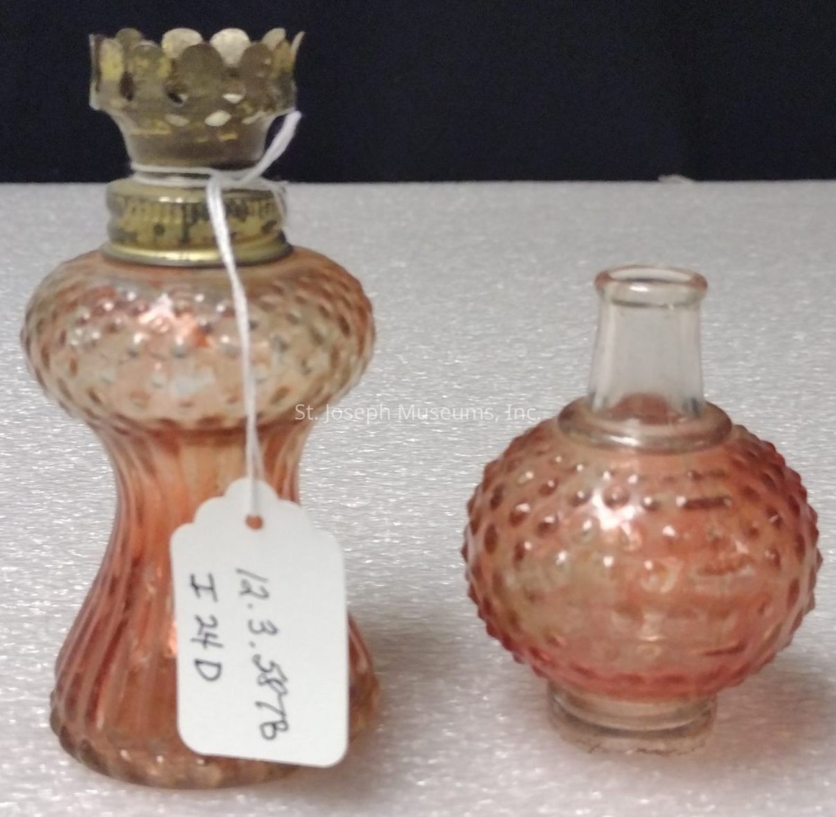 The image depicts two vintage glass bottles with a textured, hobnail design and an iridescent finish, lending them an antique aesthetic. The bottle on the left features a wider base, a narrow neck, and a decorative brass or metal crown-like cap on top. Attached to this larger bottle is a white tag with handwritten numbers and letters. The bottle on the right is smaller and rounder, with a short neck and no cap, showcasing the same hobnail design. Both bottles possess a rosy hue, shimmering under the light, placed against a textured light-colored background that contrasts with the deepened shades of the glass.