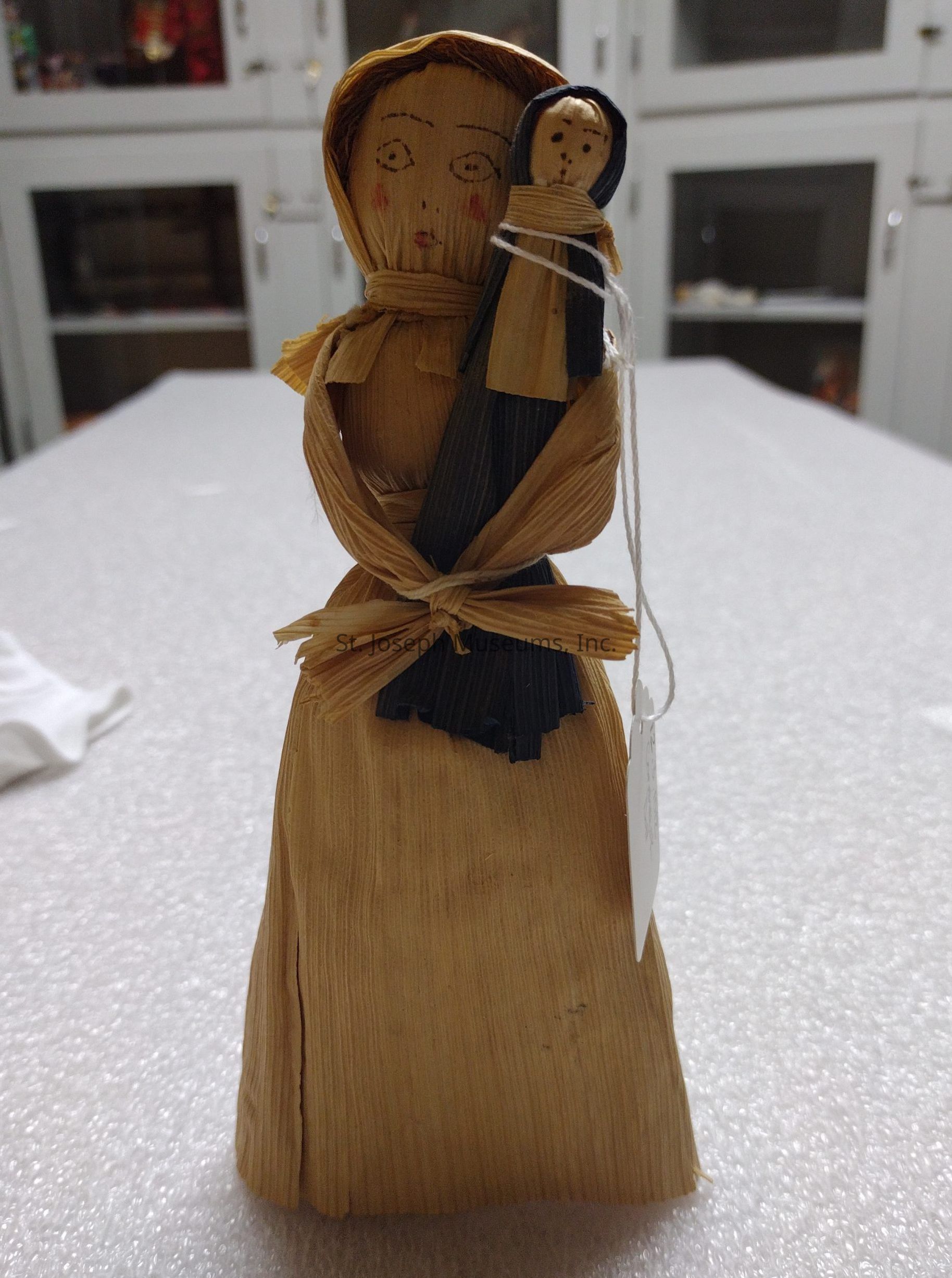 The image depicts a handcrafted figure made from corn husks, showcasing a person wearing a hooded outfit and carrying a smaller figure wrapped in the same material. The larger figure has simple facial features drawn on its face, with cheeks accentuated with a hint of color, conveying a handmade aesthetic. The smaller figure, appearing to be a child, is nestled in front of the larger one, seemingly secured by a piece of string. The background consists of white cabinetry with glass doors, suggesting the setting is a display area, while the foreground features a textured surface that contributes to the rustic and traditional feel of the scene.