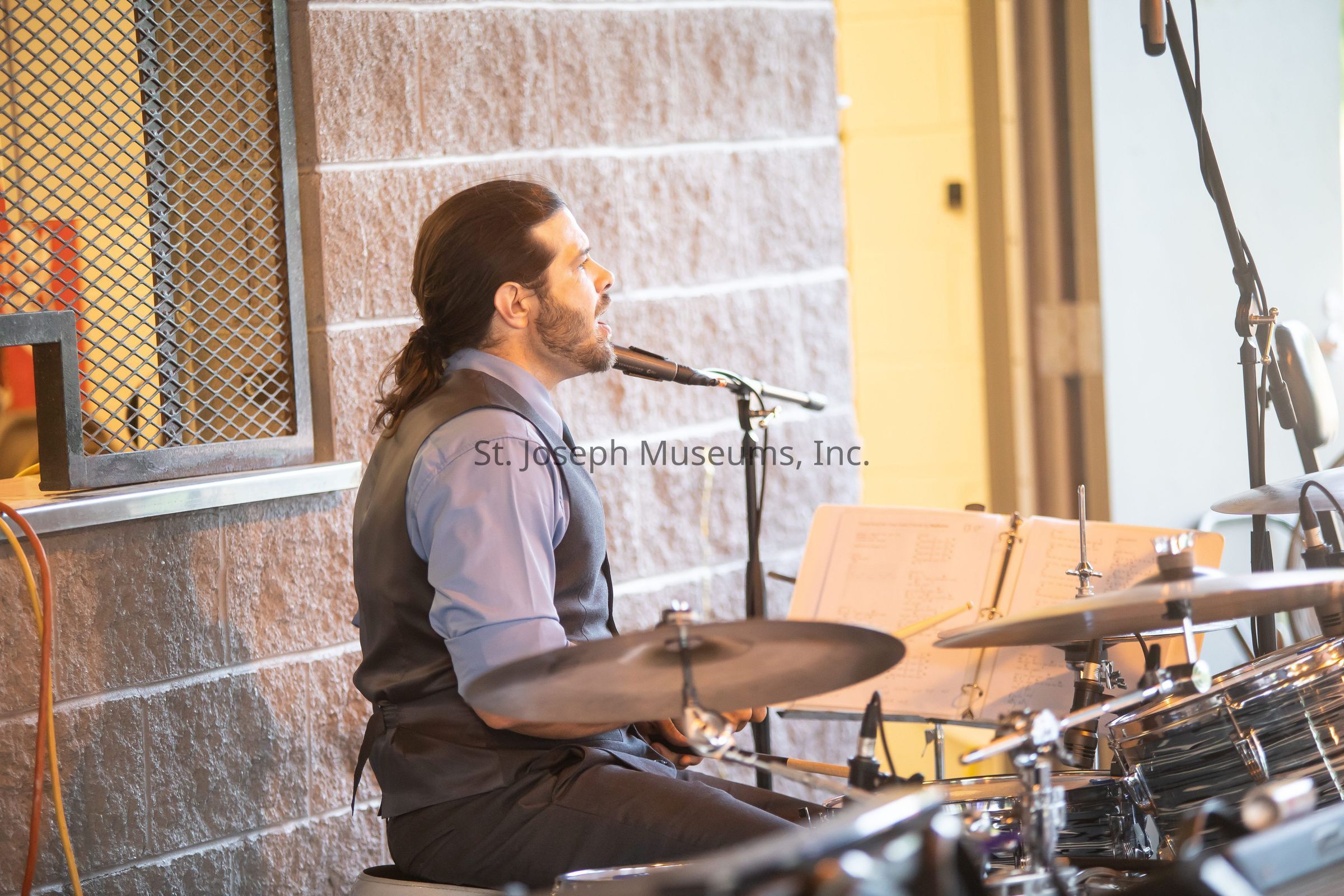 The image shows a drummer seated at a drum set, performing passionately while speaking or singing into a microphone positioned close to his mouth. He is wearing a blue dress shirt and a dark vest, with his hair pulled back, and appears focused on his performance. The drum set is equipped with various cymbals and drums, and there is a music stand displaying sheets of music in front of him. The setting is an indoor venue with a brick wall in the background, and the lighting is bright, highlighting the musician's expressions as he immerses himself in the musical moment.