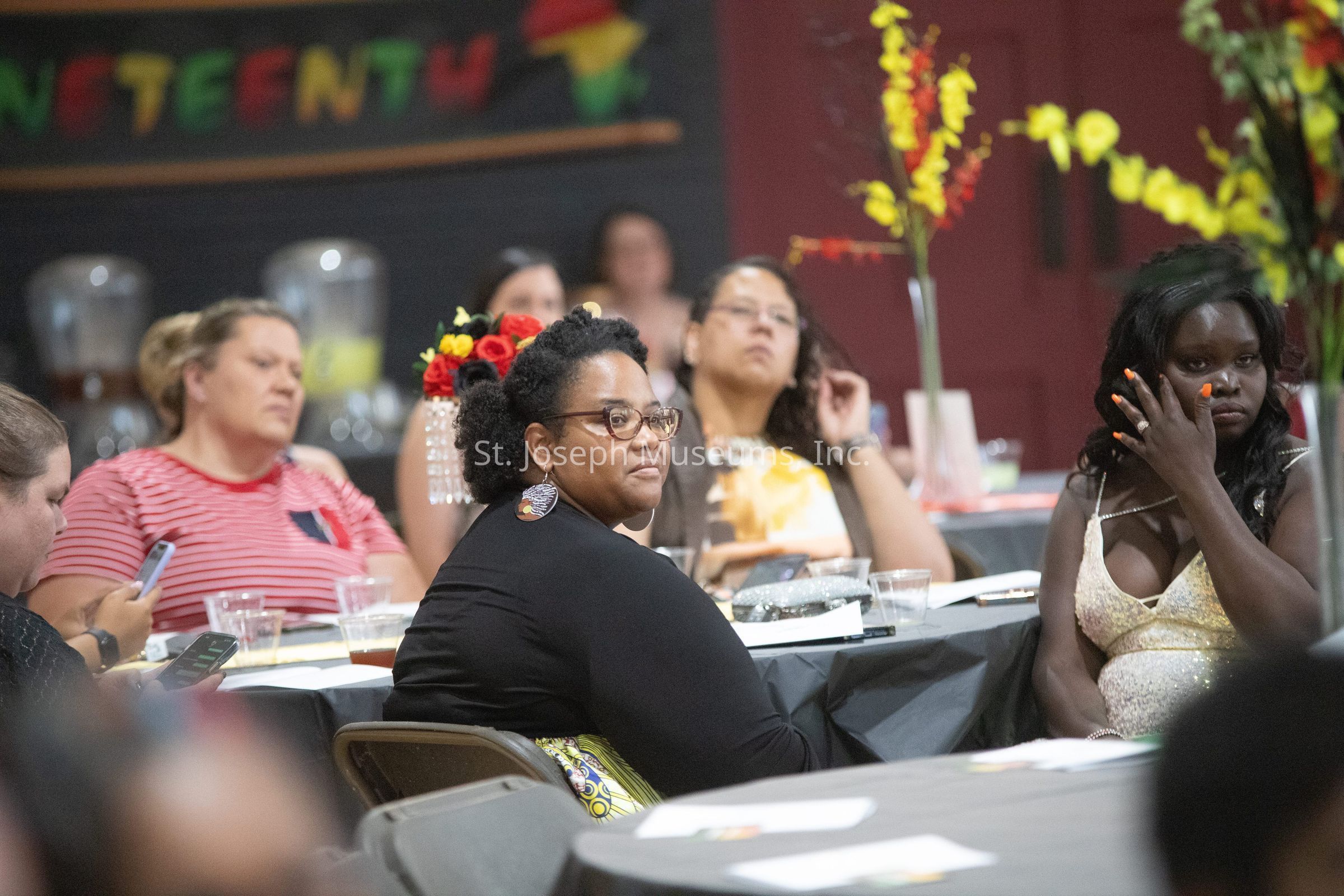 The image depicts a group of people seated around tables at an indoor event, where the background features colorful decorations and a banner partially showing the word "JUNETEENTH." The attendees, comprised of men and women of various backgrounds, are engaged in listening or watching something happening out of frame. They are sitting on folding chairs covered with black tablecloths, which bear items such as cups, notebooks, and phones. The environment is decorated with vibrant floral arrangements featuring red, yellow, and orange flowers, adding a festive atmosphere to the gathering. Some individuals are focused on their smartphones, while others look attentive to the event proceedings, reflecting a relaxed yet engaged atmosphere. The diverse assembly and the decorative theme suggest a celebration or educational event commemorating Juneteenth, a significant historical occasion in the United States.