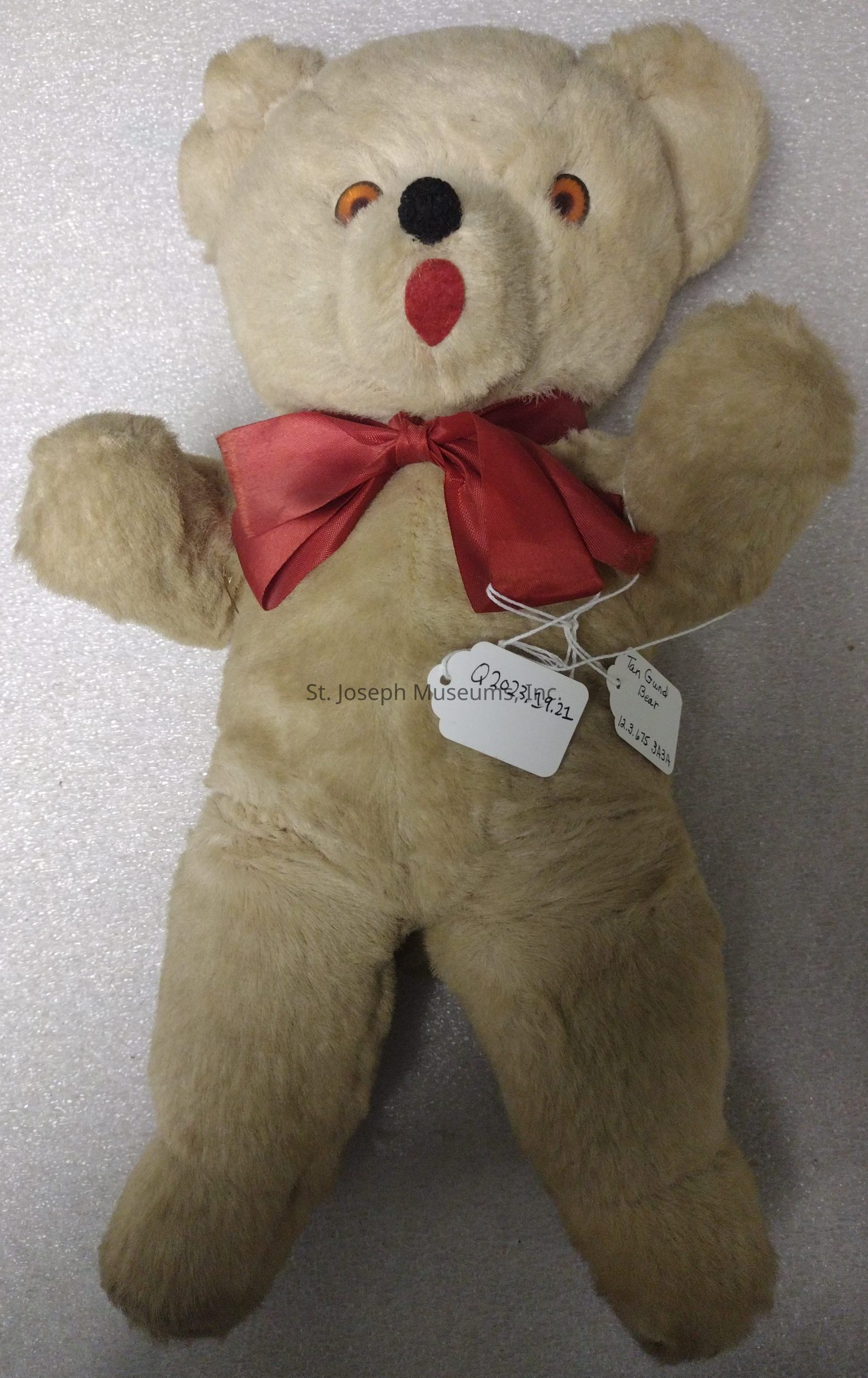 The image shows a vintage teddy bear with a light beige fur, round and fluffy, featuring a red satin bow tied around its neck, giving it a charming appearance; the bear has distinctively round eyes with a hint of orange and a small black nose which adds to its classic and enduring appeal, maintaining the timeless design beloved by collectors and children alike; attached to the bear are two tags, one labeled with "Q2023.19.21" and the other containing handwritten text "Teddy Bear Color" with additional numbers and letters, presumably for cataloging or identification purposes, indicating that this particular teddy bear may be part of a collection or museum exhibit, preserved to showcase the nostalgic essence and cultural significance of traditional stuffed animals.