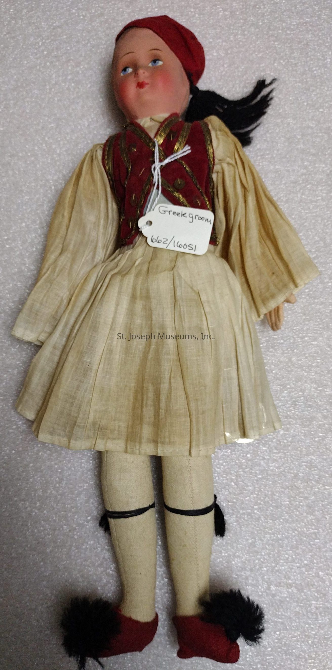 The image shows a doll dressed in traditional attire reminiscent of a Greek groom. The doll features a red cap and a matching vest with gold detailing. The main garment is a cream-colored pleated dress, which is typically associated with the traditional Greek fustanella skirt. The doll is also wearing long beige stockings, secured with black ribbons near the knees, and red shoes adorned with black pom-poms. A tag attached around the neck, labeled "Greek groom," provides additional context to the costume theme. The background is a soft, textured surface, enhancing the traditional and cultural aesthetic of the doll’s attire.