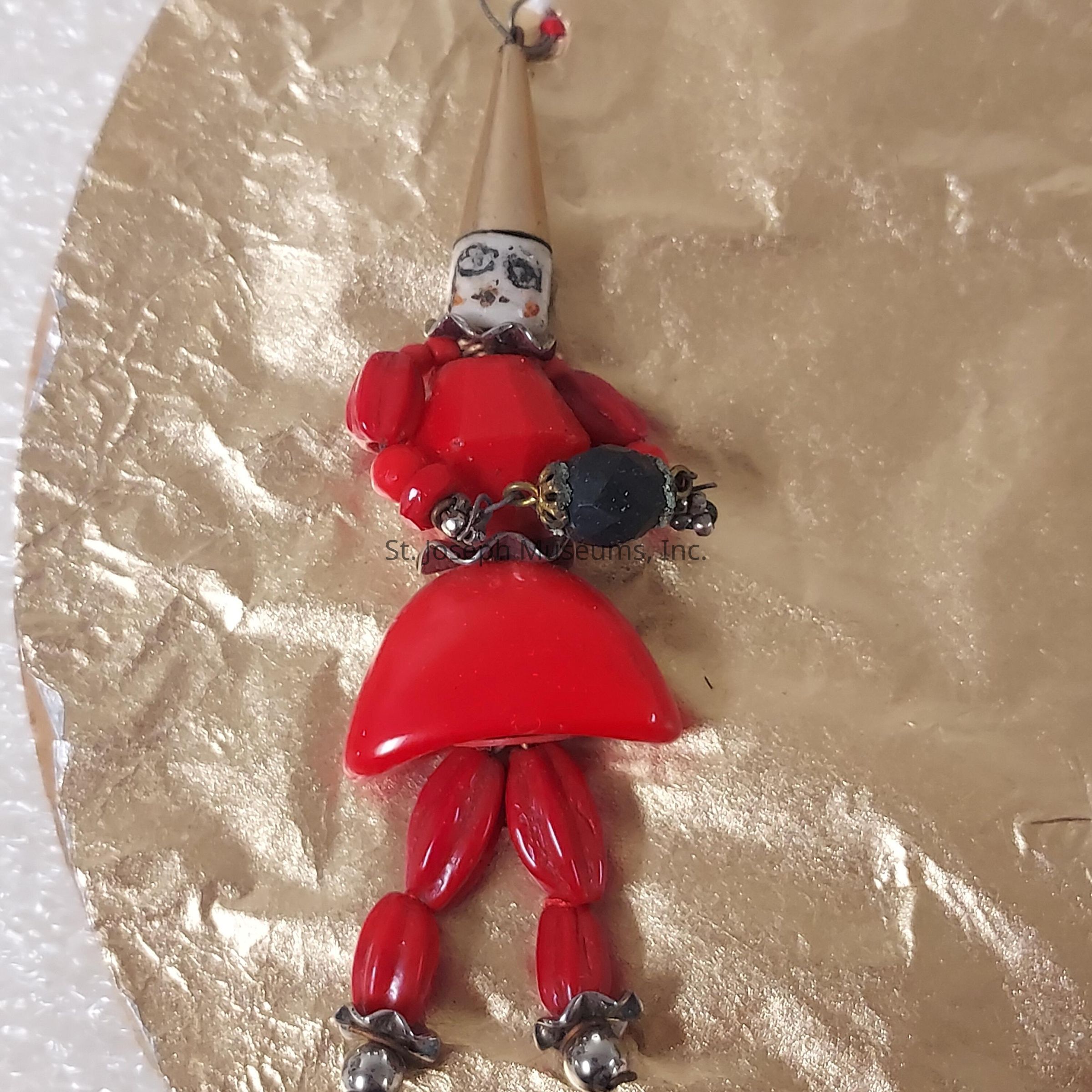 The image showcases a small, handcrafted figure made predominantly from red beads arranged to form a body, arms, and legs, with a distinctive conical headpiece. The figure is designed as a clown or jester, evident by its exaggerated features, including a painted face with black and white details. Its hands hold a spherical black object, adding a contrasting element to the predominantly red attire. The figure appears to be set against a crinkled, reflective gold background that enhances the vibrant colors of the beads, giving a festive and whimsical appearance to the entire composition.