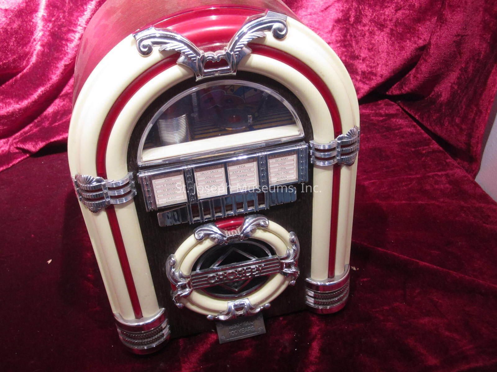 12.3.555: The image depicts a classic jukebox with a vintage design, adorned in a rich combination of cream and red colors, accentuated by chrome details and art deco elements. The jukebox features a prominent arch shape, and its front is adorned with decorative embellishments that highlight its nostalgic appeal. Below the arched glass panel, one can see rows of potential song selections, hinting at the jukebox's purpose of playing music selections. The surrounding rich red velvet backdrop adds to the dramatic and elegant presentation of the jukebox, emphasizing its status as a piece of retro art and entertainment.