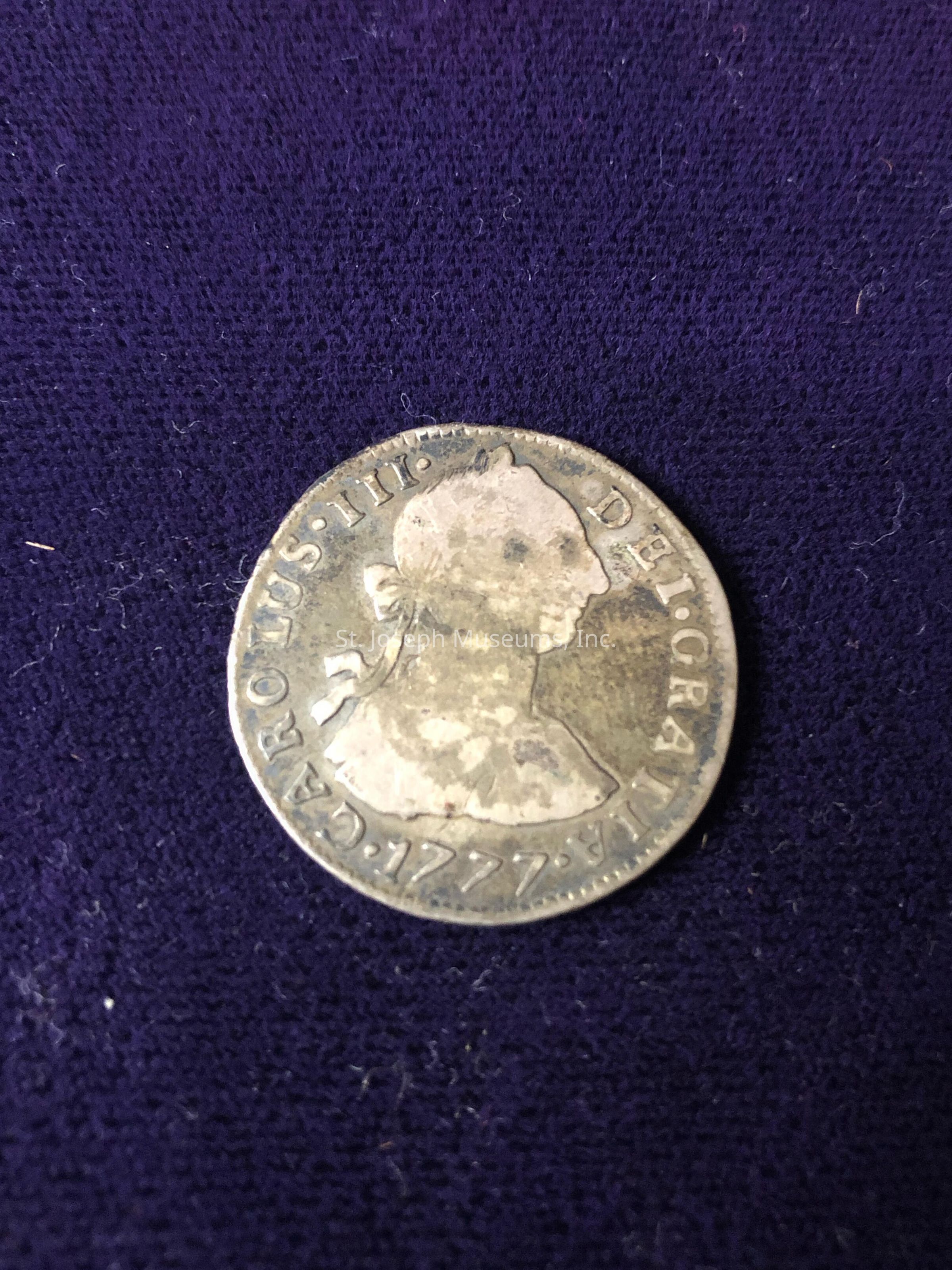 The coin features a profile of a person wearing a traditional European hairstyle from the 18th century. Around the edge of the coin, there is an inscription in Latin that reads "CAROLUS III DEI GRATIA" followed by the date "1777." The coin exhibits signs of aging, with a slightly worn and tarnished surface, indicating its historical nature. The fabric beneath the coin has a slightly fuzzy appearance, enhancing the overall vintage and antique aesthetic.