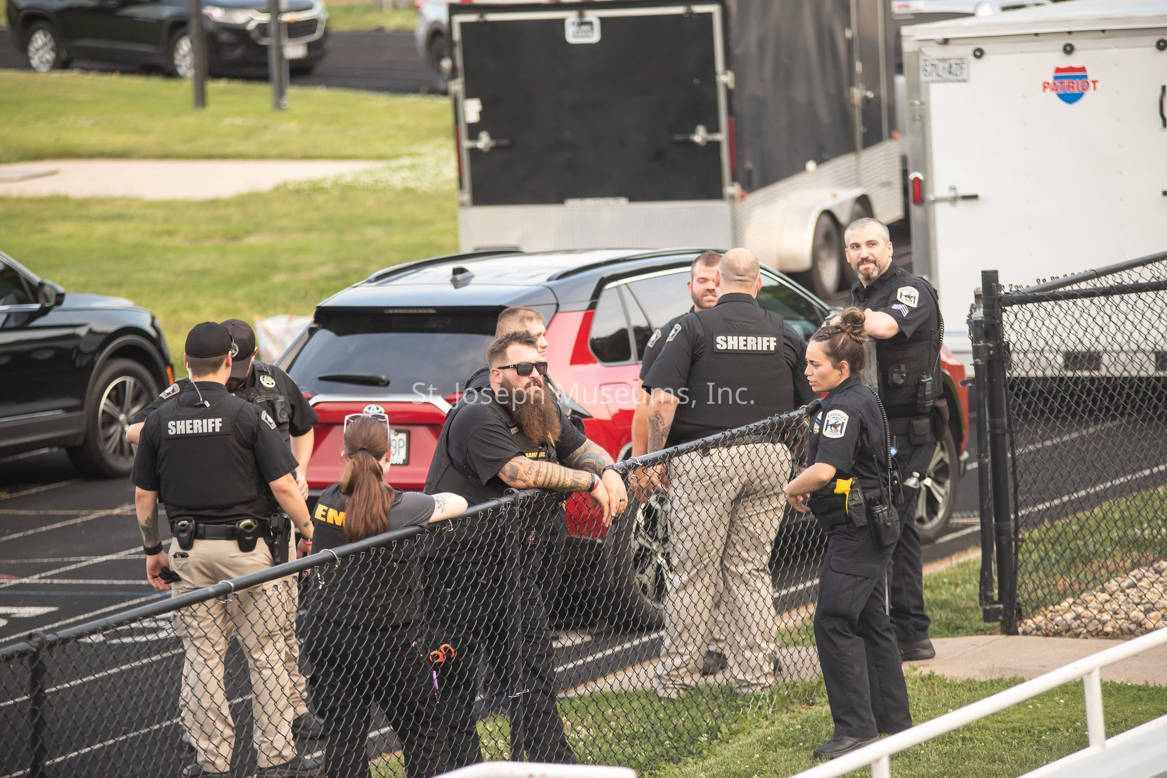 The image depicts a group of law enforcement personnel, identifiable by their uniforms and the word "SHERIFF" on their clothing, gathered near a parking area with several vehicles, including a red SUV and a large trailer in the background. Some officers appear to be engaged in conversation and appear relaxed given their postures and facial expressions. A man with a long beard, wearing sunglasses and heavily tattooed on both arms, leans casually against a chain-link fence, which separates the officers from the grassy area in front of them. Another officer, with her hair in a bun, is standing nearby with her hand on her hip, suggesting a mix of ease and professionalism. The setting suggests a non-urgent situation, possibly routine or preparatory, and the overall atmosphere conveys a sense of camaraderie among the officers. The area appears to be part of a larger, open, outdoor space with the ground and pavement visible underneath the parked vehicles.