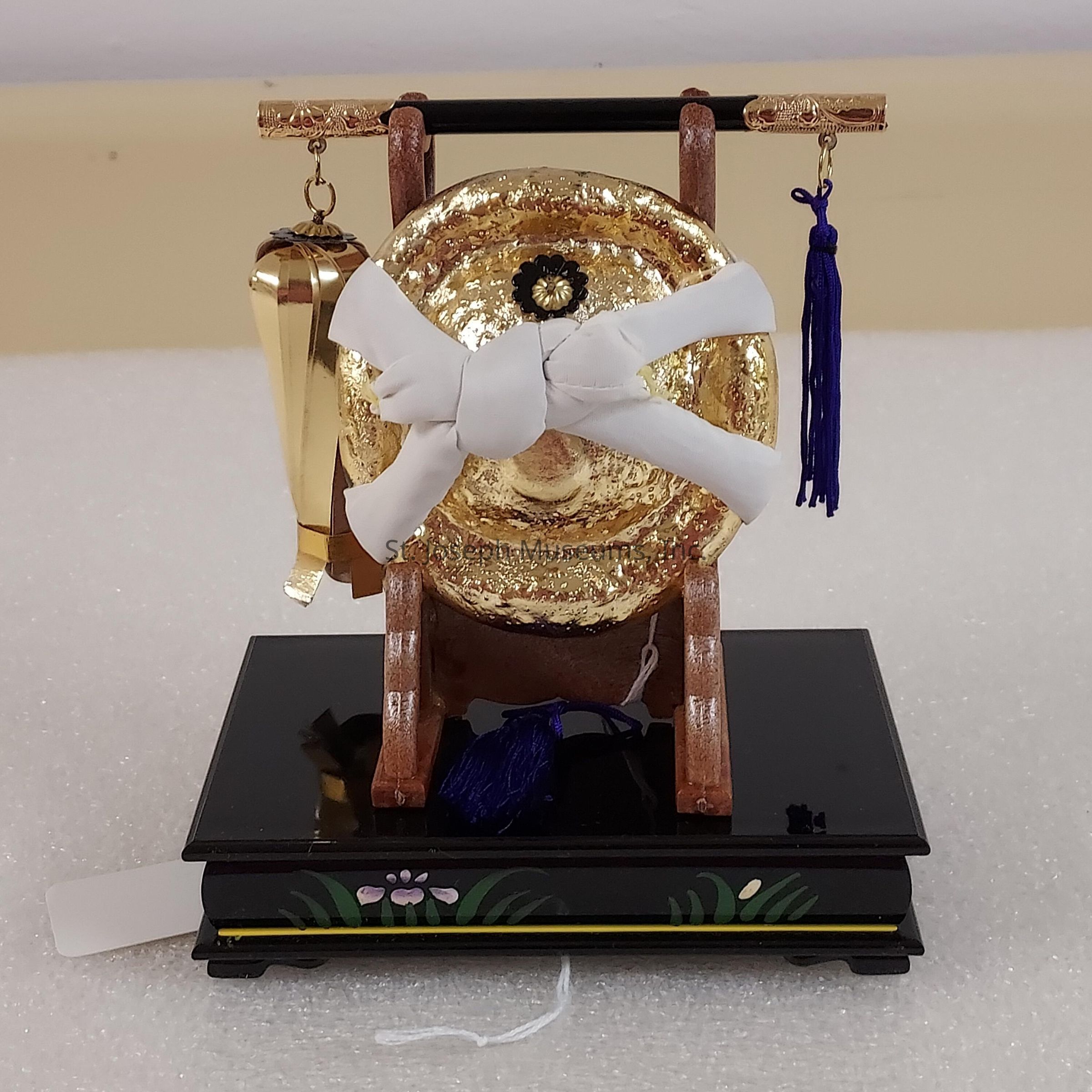 In the image, there is a decorative miniature gong displayed on a glossy black pedestal adorned with floral designs. The gong itself has a bright golden surface and is elegantly tied with a white ribbon at the center. It is supported by a wooden frame, from which two ornate tassels dangle, one in metallic gold and the other in deep purple with intricate tassel designs. The detailed craftsmanship suggests that this piece may be a ceremonial or decorative artifact, possibly used for cultural or religious purposes. The background is a soft beige, which complements the rich colors of the gong and its embellishments, making it the focal point of the composition.
