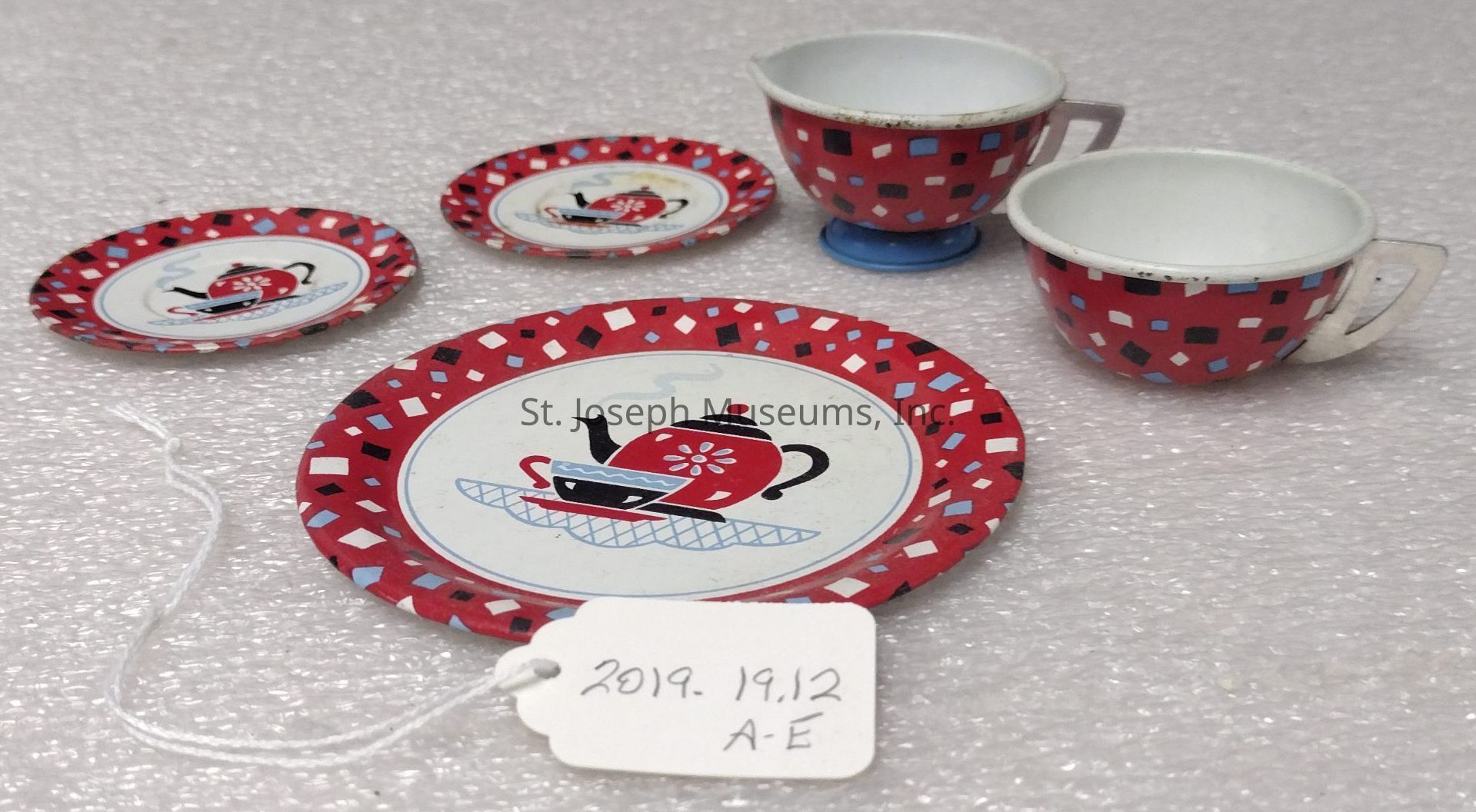 The image shows a charming vintage toy tea set consisting of two small cups and three saucers, each decorated with a playful red and white pattern featuring black and blue geometric shapes. The central design on the saucers depicts a steaming teapot and a cup on a tray, evoking a nostalgic afternoon tea setting. The saucers and cups, made of tin or metal, display a classic retro aesthetic, while the cups feature small handles and are accompanied by a tag labeled "2019.19.12 A-E," suggesting an inventory or collection reference. The setting is against a textured, light-colored surface that highlights the vivid colors and intricate details of the tea set, capturing a sense of playful nostalgia and childhood charm commonly associated with such vintage toys.