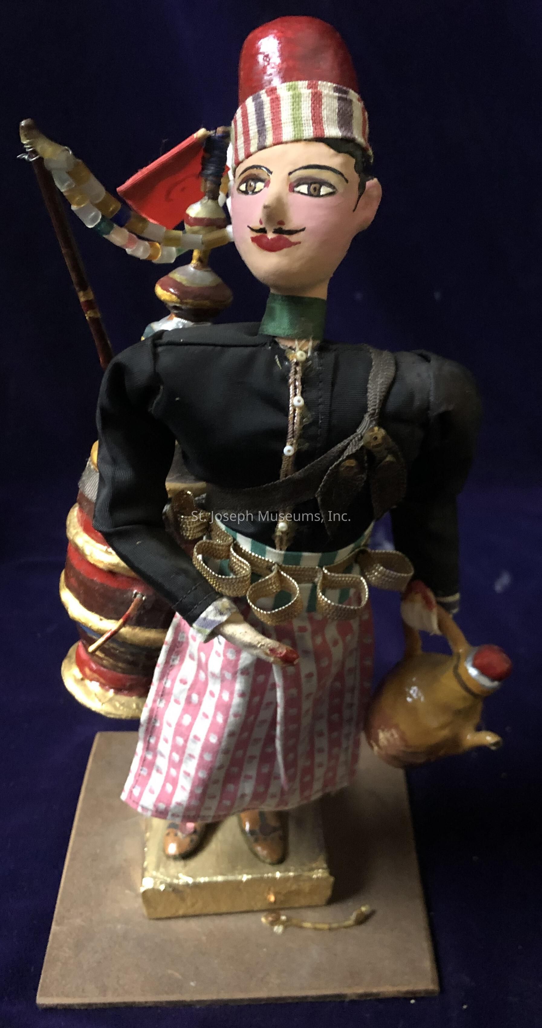 The image shows a detailed figurine of a man wearing traditional attire. He dons a striped hat and has a painted face featuring prominent eyes and a mustache. His outfit consists of a black jacket, a patterned red and white skirt, and a belt with multiple loops. He carries a kettle-like container in one hand, and on his back, there appears to be a set of pipes or instruments with colorful accents. The figure stands on a base, and the background is a deep, dark color that contrasts with the vibrant colors of the figurine, enhancing its intricate design.