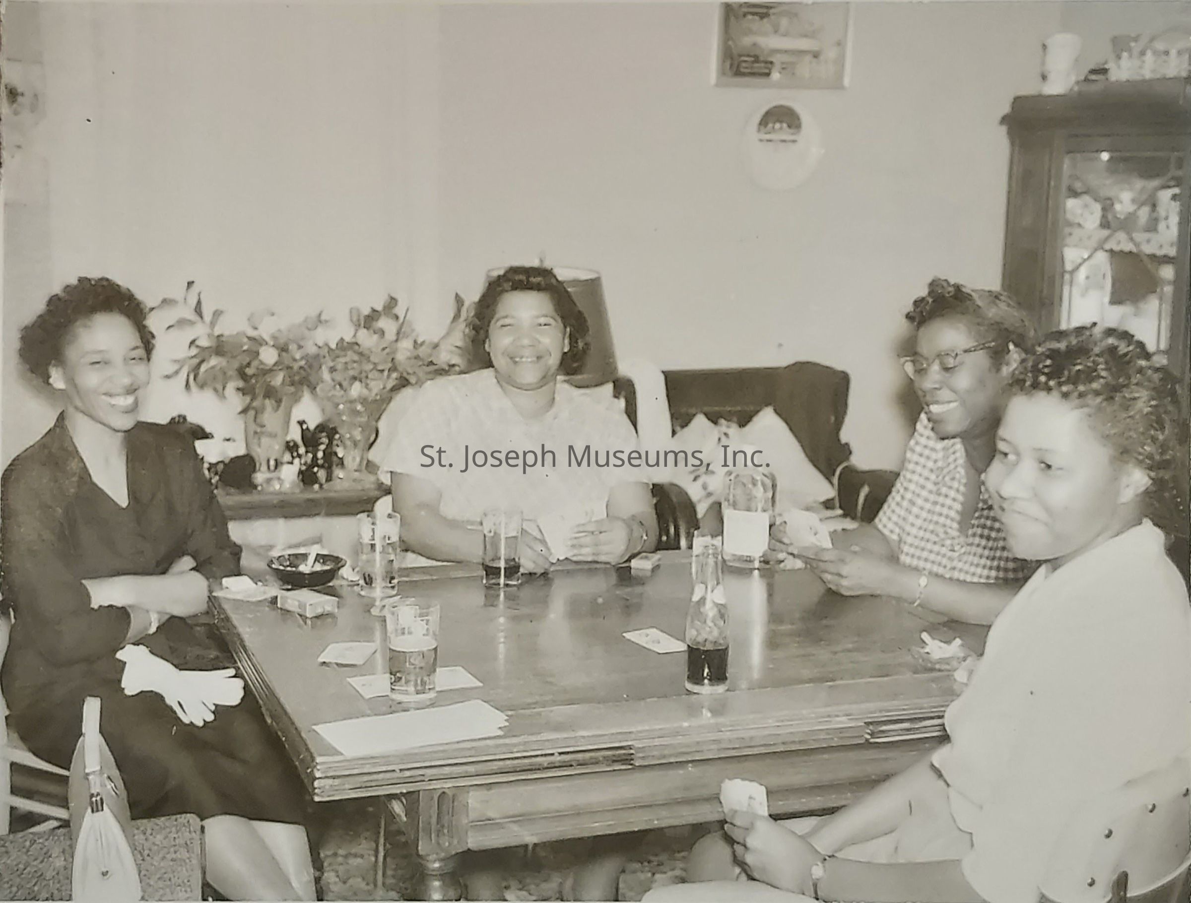 In the sepia-toned image, four women are seated around a wooden table in a cozy, warmly lit room, each engaged in playing a card game. They appear to be enjoying a relaxed social gathering, as evidenced by their joyful expressions and relaxed postures. The tabletop holds several glasses, some with drinks, while a few playing cards and small items are scattered about, suggesting a casual, friendly card game in progress. Each woman is dressed in mid-20th century attire, radiating a sense of camaraderie and leisure. In the background, a large bouquet of flowers adds a touch of elegance, and a cabinet displays various decorative items, contributing to the inviting atmosphere of the room. The scene captures a moment of shared happiness and connection among the women, emphasizing the warmth and simplicity of such social interactions.