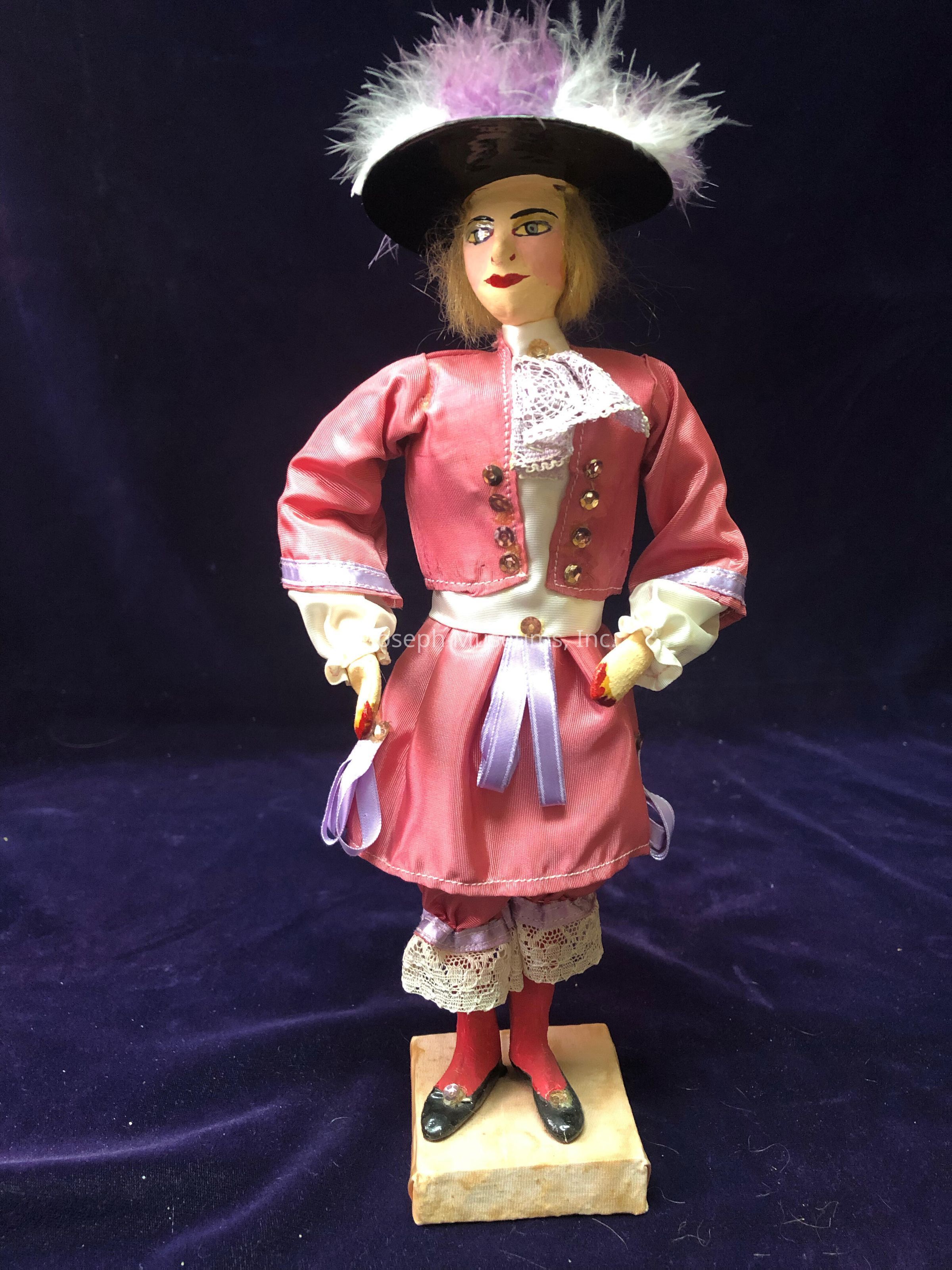 The image showcases a detailed figurine dressed in period-style clothing, featuring a red and white ensemble with a matching jacket and skirt accented by lace and ribbon details. The doll wears a pair of red stockings and black shoes, and the sleeves of the jacket are adorned with lace cuffs. Atop its head sits a wide-brimmed black hat embellished with white and purple feathers. The figure stands confidently on a small square pedestal, set against a rich, dark velvet background that enhances its vibrant colors and intricate design.