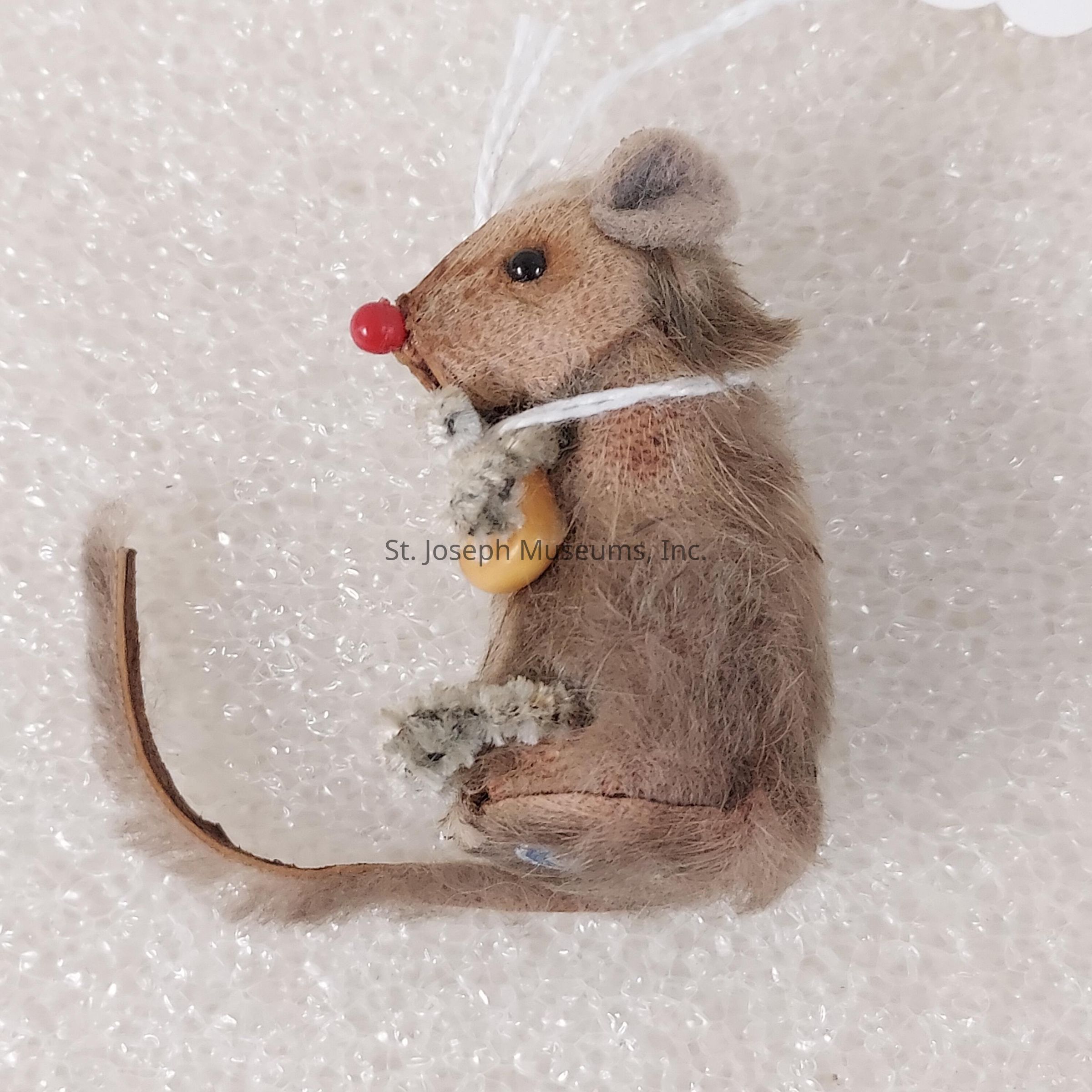 The image depicts a small, realistic-looking mouse figurine set against a textured, white background. The mouse has a soft, furry appearance with brown fur and a lighter colored underbelly. It features detailed facial elements like a tiny, shiny black eye and a red nose, giving it a charming and lifelike expression. The mouse is sitting in an upright position, holding a small object that appears to be an amber bead or seed. A string is looped over the mouse’s head, suggesting it is an ornament or decoration. Its tail is long and slender, curving naturally, contributing to its realistic representation. The textured background heightens the focus on the intricately designed figurine.