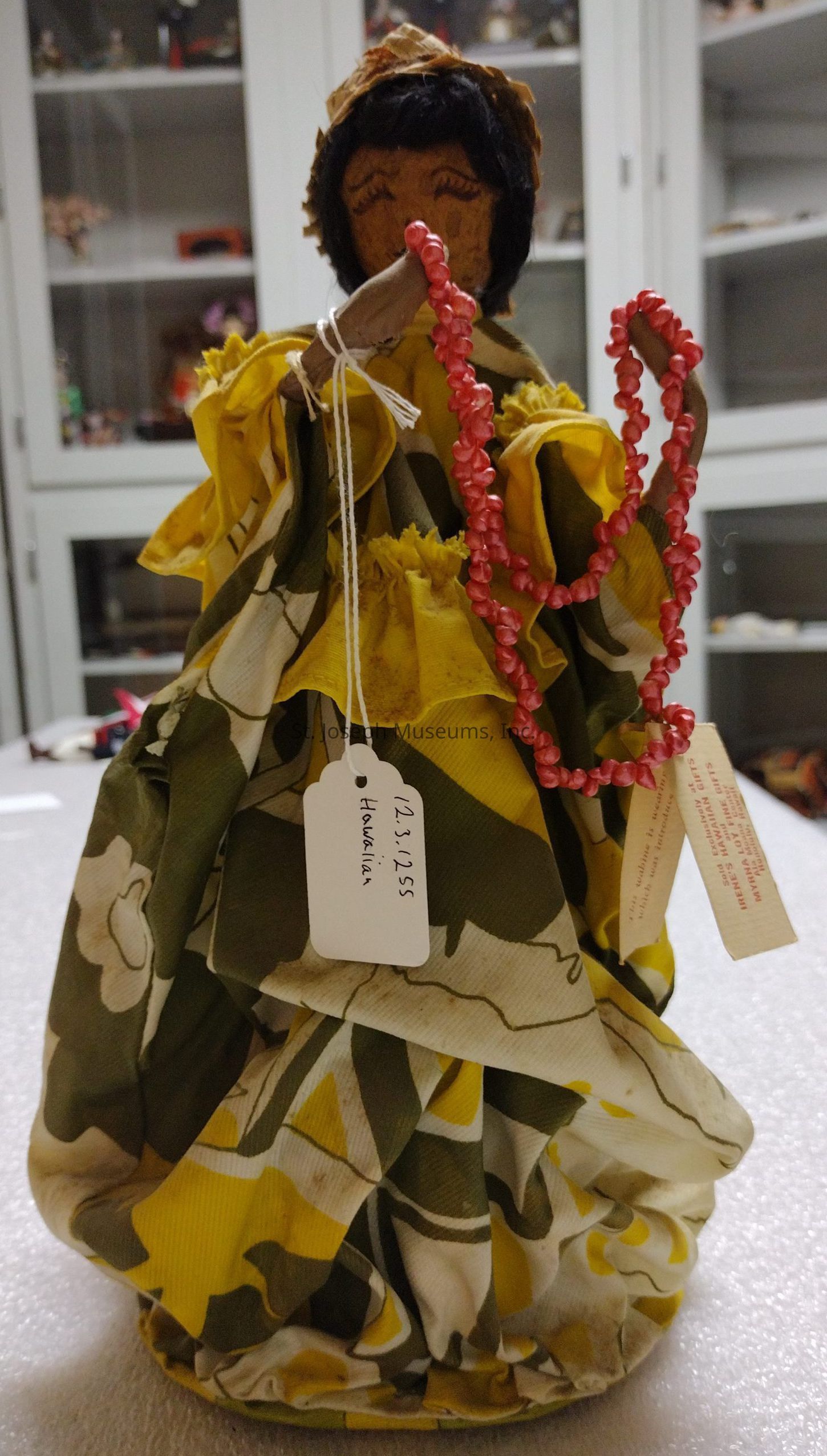 The image depicts a handmade cloth doll, which has a simple facial expression with closed eyes and black hair. The doll is adorned with a multicolored dress featuring vibrant yellow, green, and white patterns, which give it a traditional yet colorful appearance. Around the neck hangs a red beaded necklace that adds a charming touch. Attached to the doll are white paper tags with handwritten text, including the date "12.3.1955" and the word "Hawaiian," suggesting cultural significance and origin. The background shows a cabinet with various objects, adding depth to the setting.