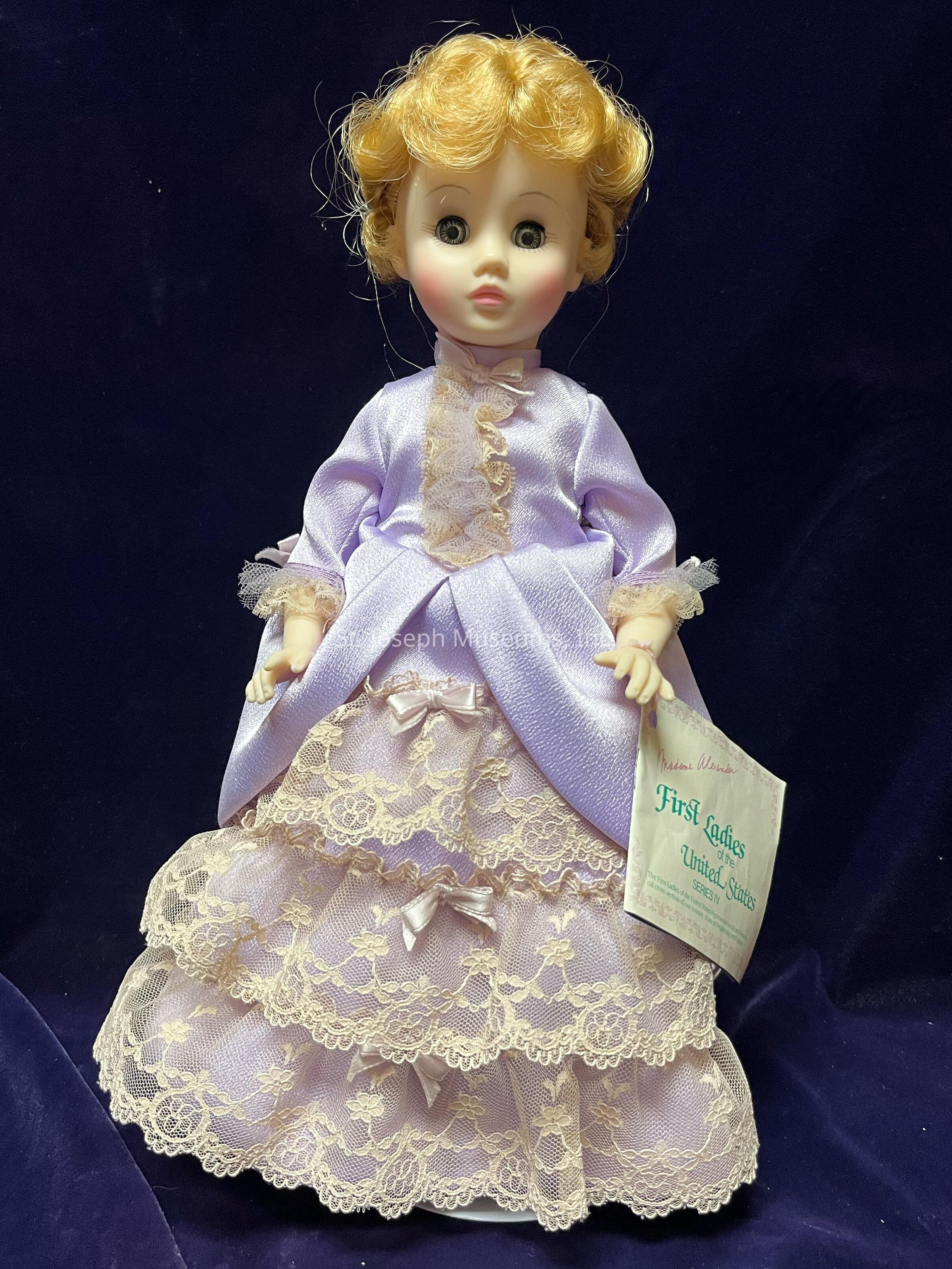 The image features a porcelain doll with curly blonde hair dressed in an elegant lavender outfit. The dress highlights intricate details with a lace-trimmed bodice and layered lace skirt adorned with small bows. The doll is in the likeness of Lucretia Garfield, First Lady and wife to James A. Garfield.