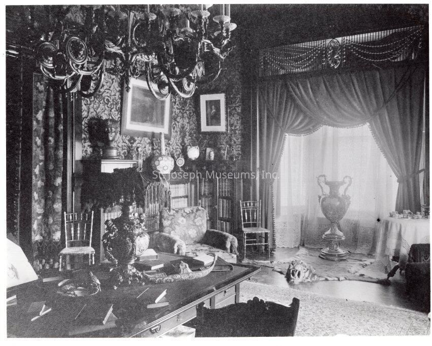 Tootle Mansion, Library, c. 1900: A black-and-white photograph of the Tootle Mansion Library around 1900 depicts a room filled with elegant furnishings and intricate decorations. A large, ornate chandelier hangs from the ceiling, casting light on a richly carved table surrounded by decorative items and a plush sofa. Behind the sofa are detailed bookcases lined with volumes, and the walls are adorned with patterned wallpaper and framed artwork. In the background, heavy drapes frame a large window, allowing natural light to filter through, highlighting a grand vase sitting on a tigerskin rug.