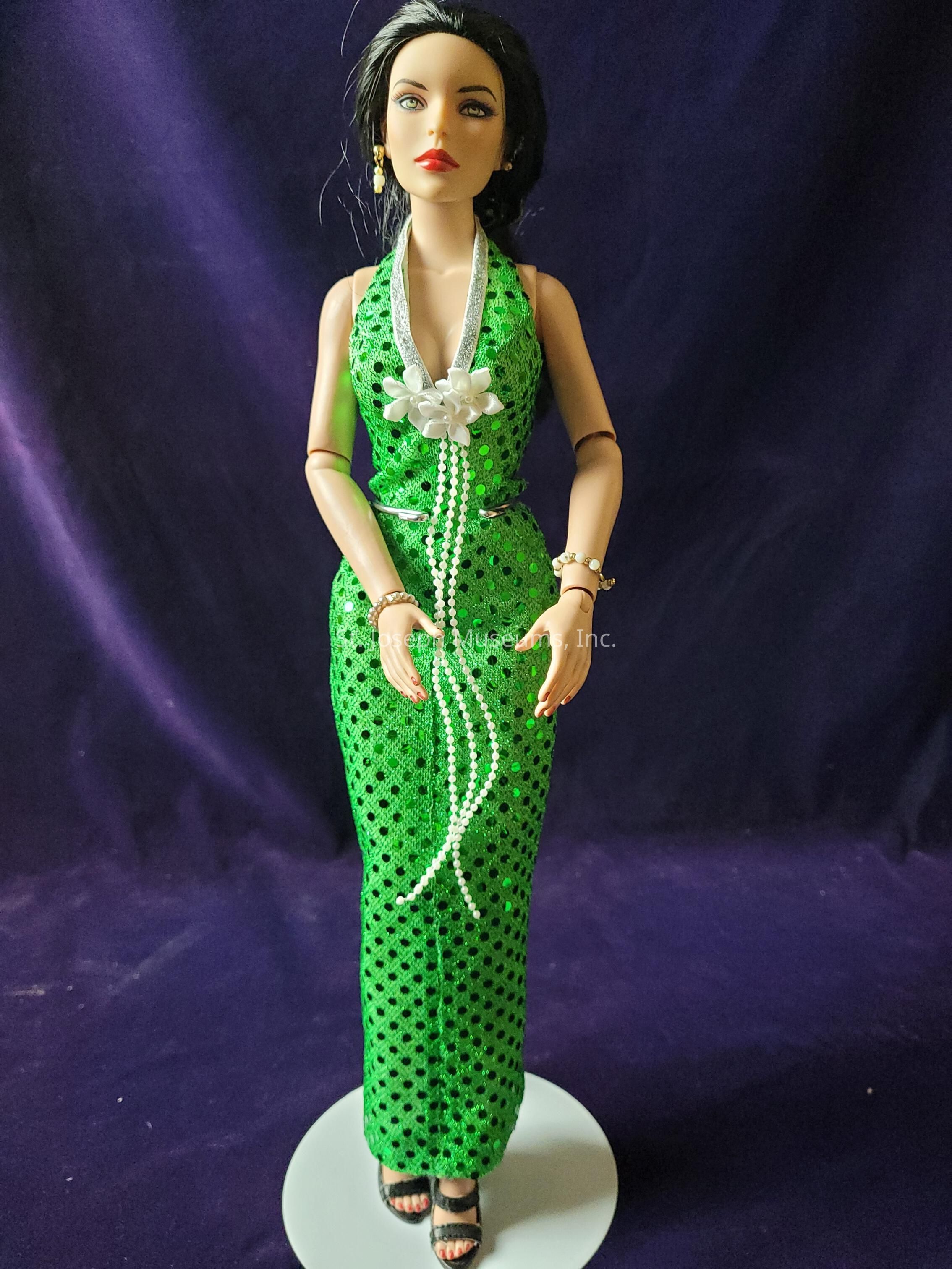 The image features a doll standing against a deep purple background, dressed in a striking green sequined gown with a halter neckline and a detailed pattern of black dots. The dress is elegantly adorned with a white floral accent at the neckline, from which pearl-like strands cascade down the front, adding a touch of sophistication. The doll has long, dark hair styled gracefully, with prominent red lips and defined facial features. She accessorizes with a delicate gold bracelet and matching earrings, as well as black strappy sandals that complete her glamorous ensemble. The doll is positioned on a white circular stand, elevating her poised stance. The overall composition of the image highlights the contrast between the vibrant green of the dress and the rich purple background, emphasizing the doll's elegant and stylish appearance.