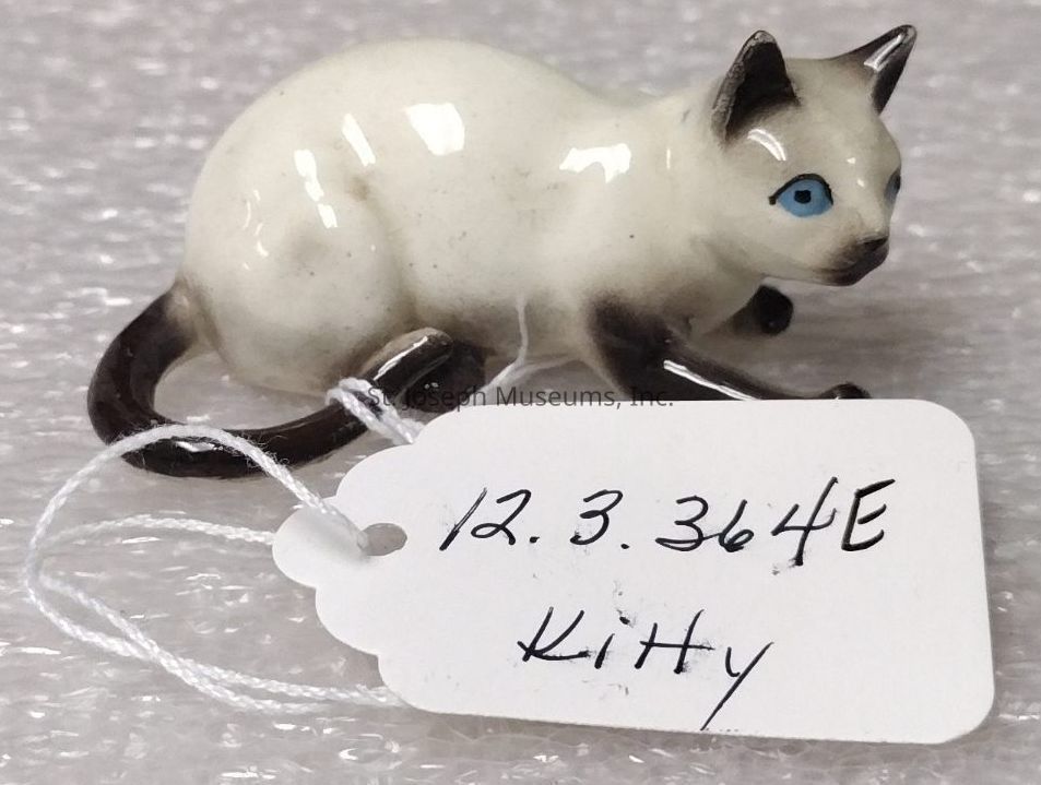 The image shows a small ceramic figurine of a cat, with a glossy surface featuring shades of white and dark accents on its ears, paws, and tail. The figurine is designed with bright blue eyes and stands in a crouching position, displaying fine details that reflect careful craftsmanship. Attached to the cat is a white string tag with handwritten text, displaying the numbers "12.3.364E" and the word "Kitty." Positioned on a textured, light-colored background, the ceramic piece reflects light, highlighting its smooth finish and artistic attributes.