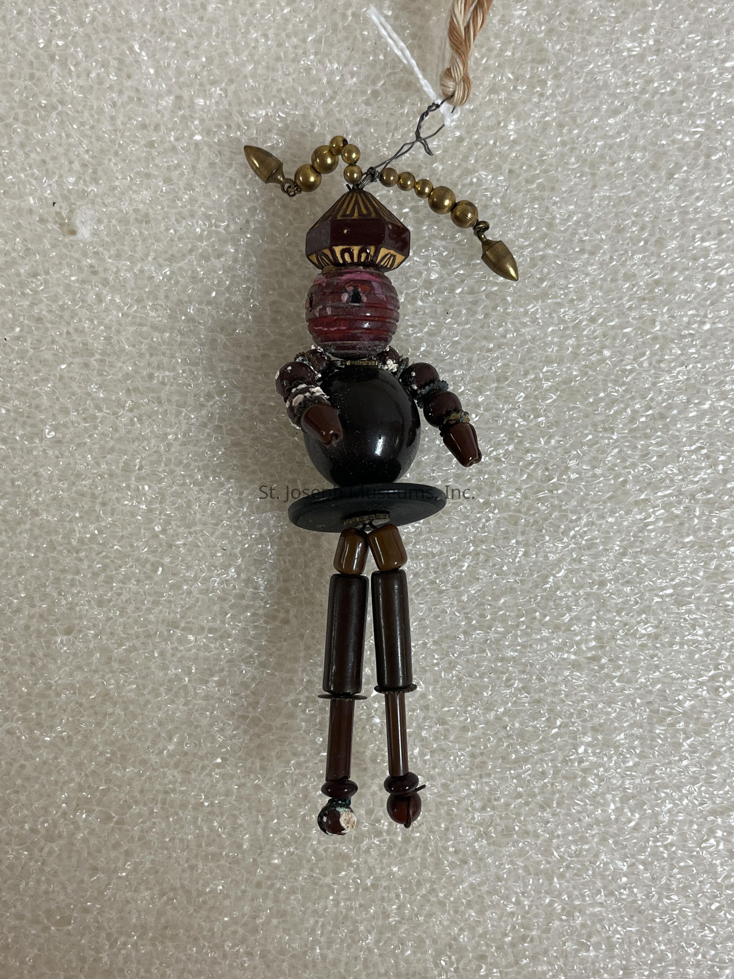 The image depicts a beaded pendant in the shape of a small humanoid figure, crafted from a variety of beads in shades of brown, black, and gold. The figure's head is represented by a large, stacked red and gold bead that resembles a stylized face. The body is a glossy black bead, while the arms and legs are composed of elongated beads, providing a charming, jointed appearance. The top of the figure is adorned with a strand of golden beads connecting to a loop, suggesting it is designed to be hung or worn. The background is a textured surface, which appears to be a type of packaging material, providing a neutral backdrop that accentuates the intricate detailing and varied colors of the pendant.