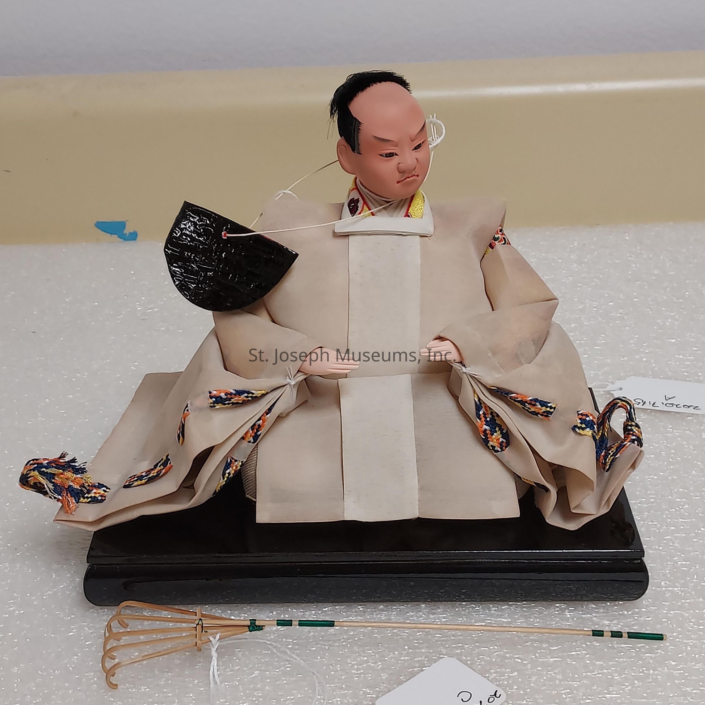 The image shows a traditional Japanese doll, meticulously crafted and dressed in a detailed, multi-layered kimono, sitting on a black platform. The kimono's outer layer is an elegant shade of beige with intricate, brightly colored patterns lining the sleeves and edges, adding a vibrant contrast. The doll's face is expressive, with sharply painted features, giving it a lifelike appearance. It holds a small black fan in one hand, secured by a string, and a delicate, ornamental rake-like object lies in front, possibly symbolizing cultural or historical significance. The background is neutral, enhancing the doll's prominence, and small tags are visible, likely indicating information about the item, further emphasizing the doll's artistic and possibly historical value.