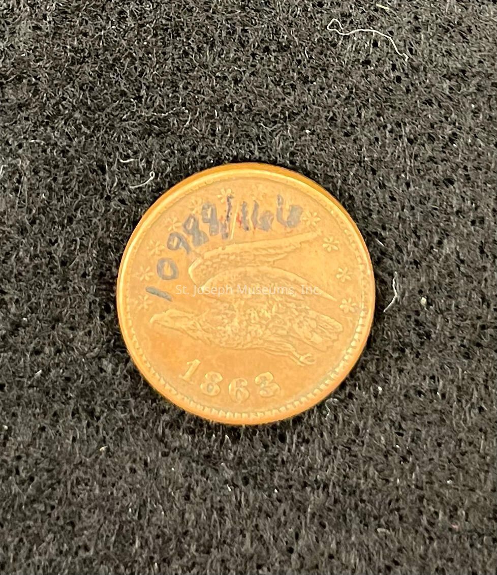The image shows a 1863 penny. The coin is a rich golden-brown color typical of old copper coins, featuring an engraved image of an eagle in flight with outstretched wings, surrounded by small stars. The year "1863" is prominently displayed at the bottom of the coin, while the top appears to be marked with some numbers or letters that seem to have been written by hand, adding an unusual and distinctive element to the antique coin's appearance. The textured background creates a contrast that highlights the historical coin, emphasizing its details and worn engravings, offering a glimpse into the numismatic history and making it a point of interest for collectors and enthusiasts alike who appreciate historical artifacts.