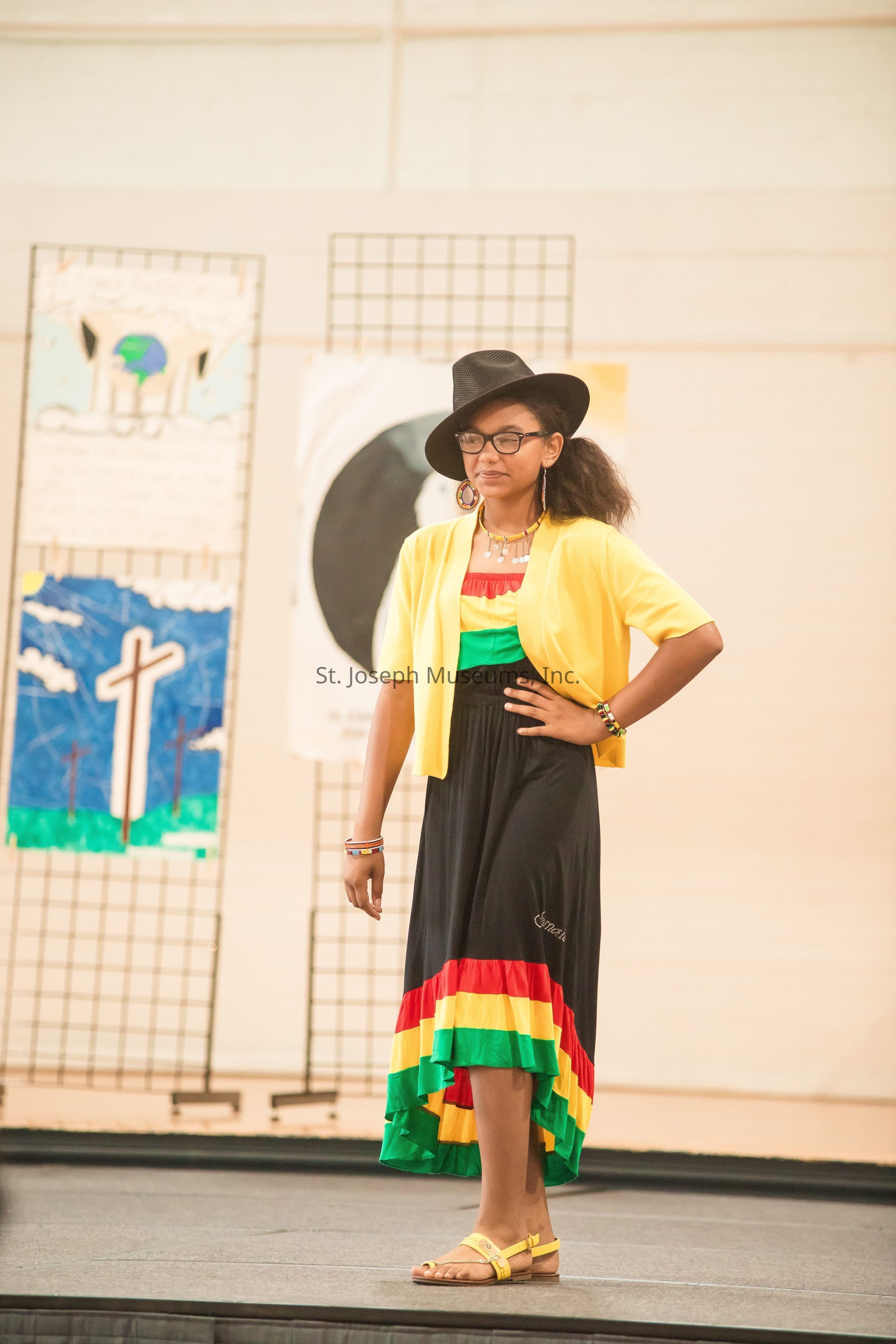 A young girl is confidently standing on a stage, dressed in a vibrant outfit consisting of a black dress with bright red, yellow, and green stripes at the hem, paired with a yellow cardigan. She accessorizes with a black wide-brimmed hat, glasses, and colorful bracelets, as well as large hoop earrings. Her pose is stylish and poised, with one hand on her hip, conveying a sense of confidence and pride. Behind her, artworks are displayed on metal grids, one featuring a cross and another with an abstract design. The setting suggests a cultural or artistic event, highlighting creativity and expression.
