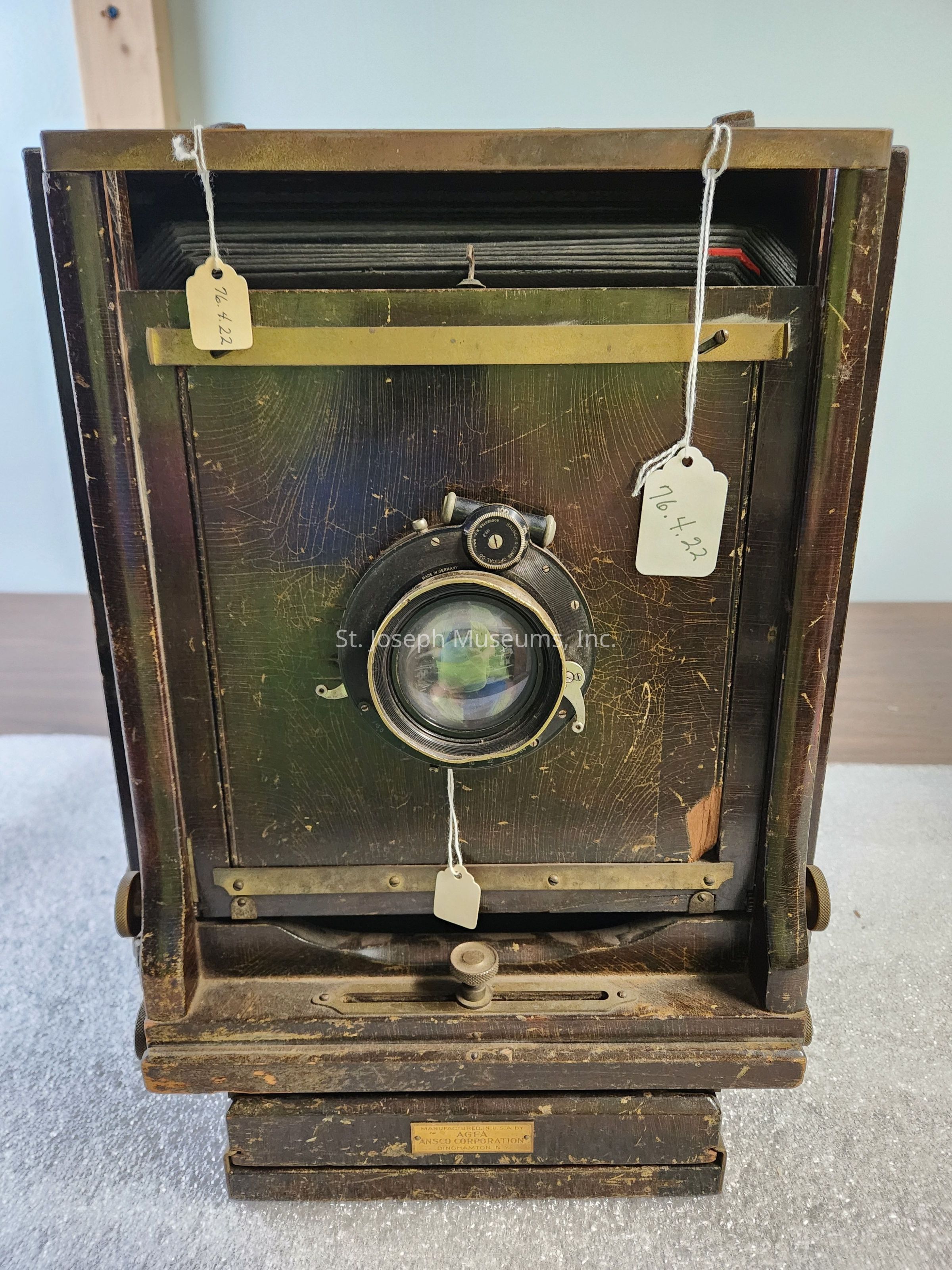 The image displays a large, vintage wooden camera with a lens in the center surrounded by a brass and wood frame showing signs of age and wear, evident from its scuffed surfaces and cracked paint. The camera’s structure is sturdy, typical of early photography equipment designed for stability and precise alignment, showcasing intricate mechanisms and levers likely used to adjust focus or aperture settings. Three small tags attached to the camera with string display numerical details, possibly for identification or cataloging purposes, hinting at its historical significance or use in a collection. This piece, with its classic, ornate design and patina, embodies the craftsmanship and technological innovation of its era, likely capturing a sense of nostalgia for photography enthusiasts and historians, as it stands as a representation of the evolution of photographic technology from cumbersome, manually operated devices to modern, digital equipment.