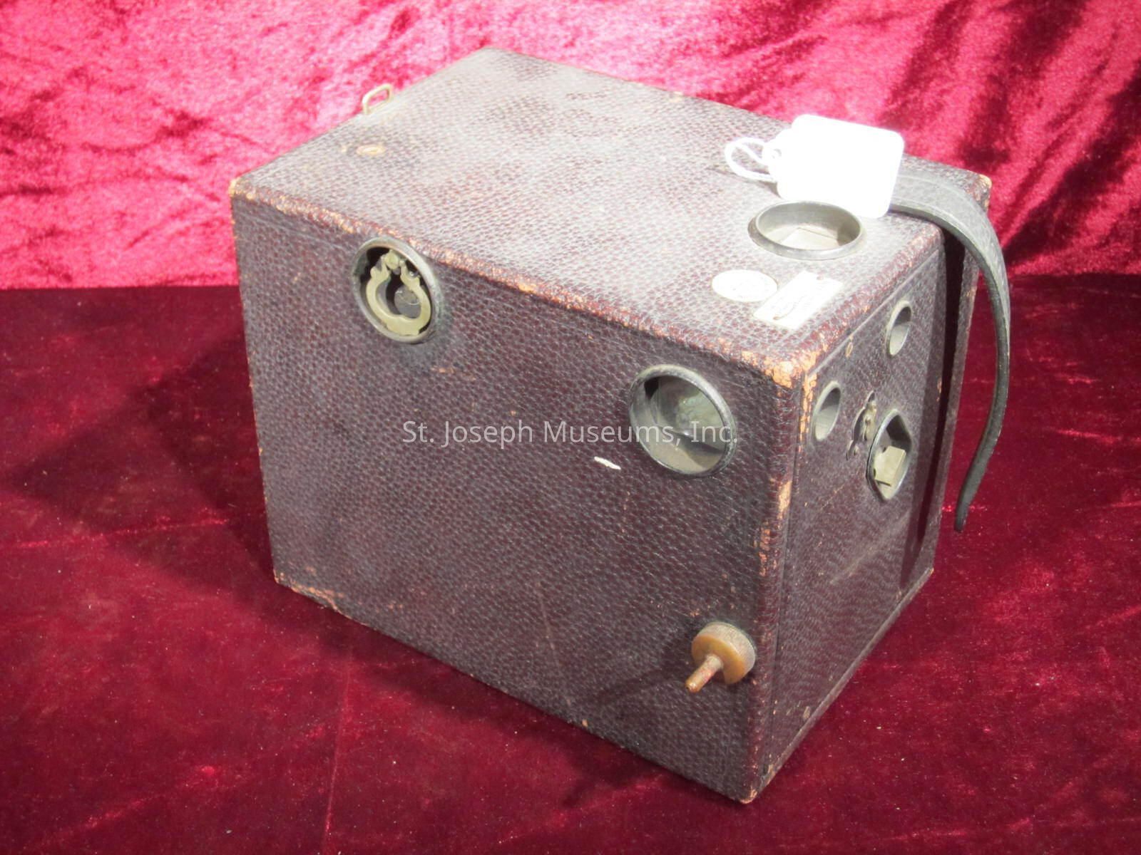 The image shows a vintage box camera placed against a rich, red velvet backdrop. The camera has a rectangular shape with a textured, dark brown covering and several circular dials and openings. It features a simple design, typical of early photographic equipment, with a worn appearance that speaks to its age and use. On one side, there is a handle, possibly made of leather, attached to the camera, and a tag hangs from a string, suggesting it may be part of a collection or for sale. The overall scene conveys a sense of historical nostalgia and a glimpse into the evolution of photography technology.