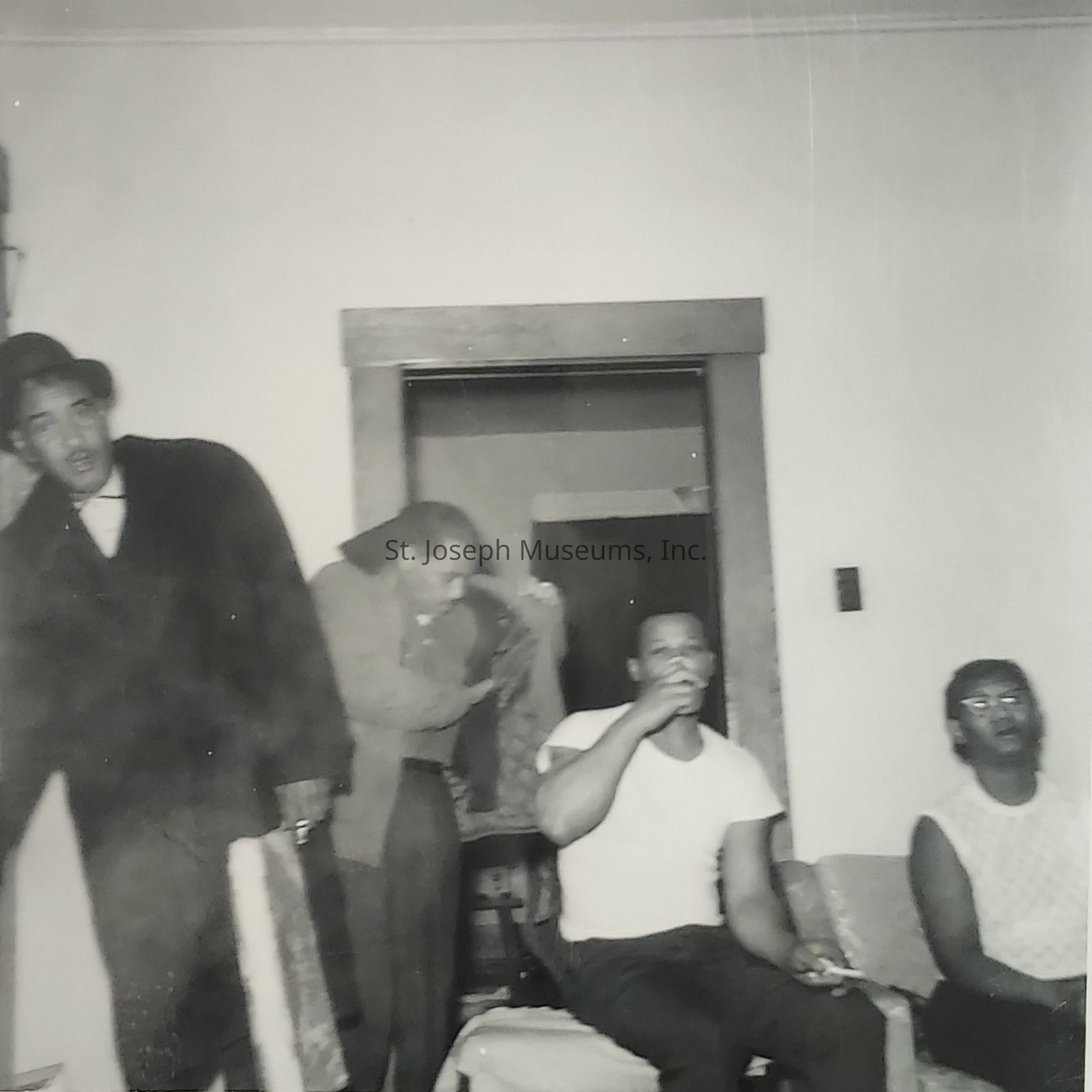 The black-and-white photograph depicts four individuals in a modest indoor setting, likely a living room. On the left, a man wearing a long black coat and a hat holds the door, showing a casual demeanor. Next to him, another man appears to be either entering or adjusting his jacket, caught mid-motion. To the right, a man in a white T-shirt and dark pants sits on a couch, seemingly engaged in thought or conversation while holding a cigarette. Beside him, another person, also seated, wears a patterned sleeveless top and glasses, looking toward the camera. The image captures a candid, everyday moment, conveying a sense of familiarity and social interaction among the group.